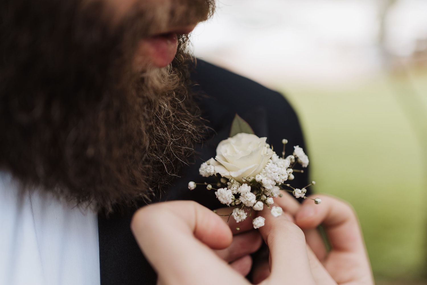 A white rose and baby's breath boutonniere being pinned onto a suit jacket with a visible beard.
