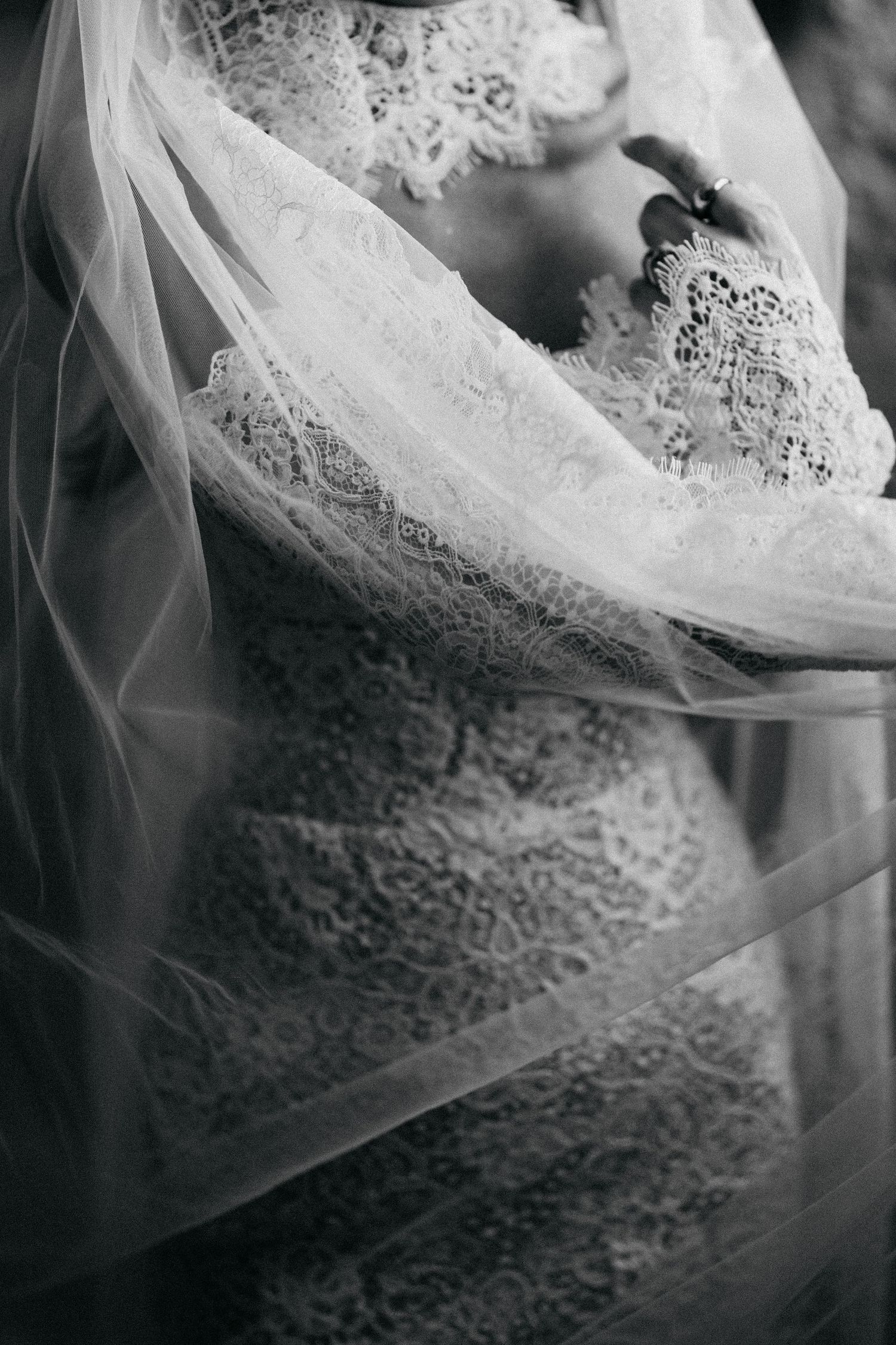 Black and white detail of a lace wedding dress with sheer long sleeves and a flowing tulle veil draped over the bride.