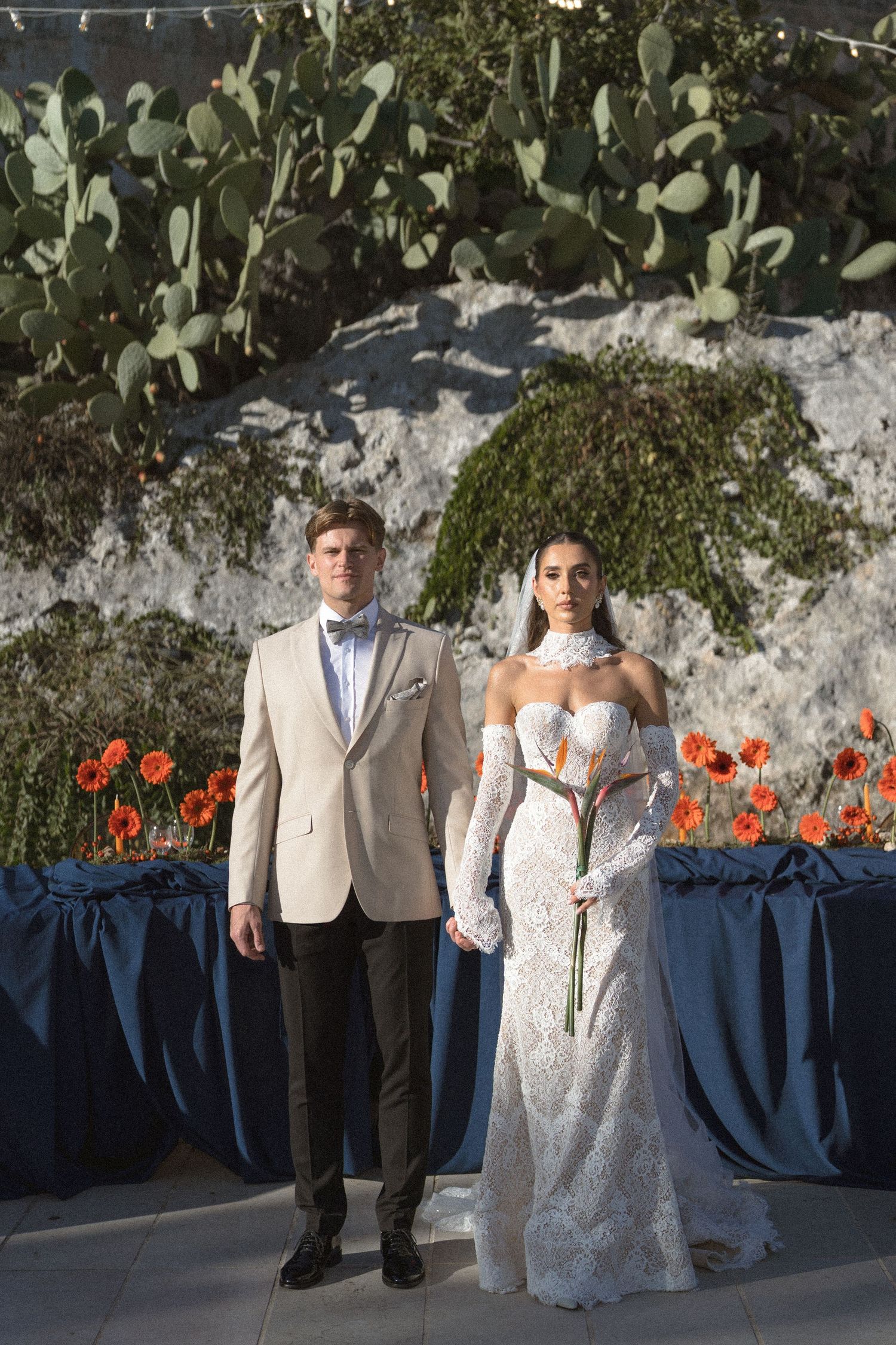 Bride in a lace gown holding an orange bouquet stands beside a groom in a beige tuxedo jacket and bow tie near cacti.