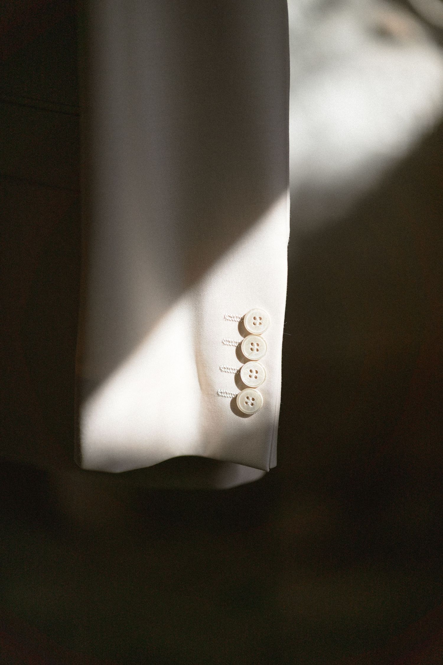 Sunlight and shadow fall across the cuff of a white suit jacket sleeve showing four buttons with contrasting white stitching.