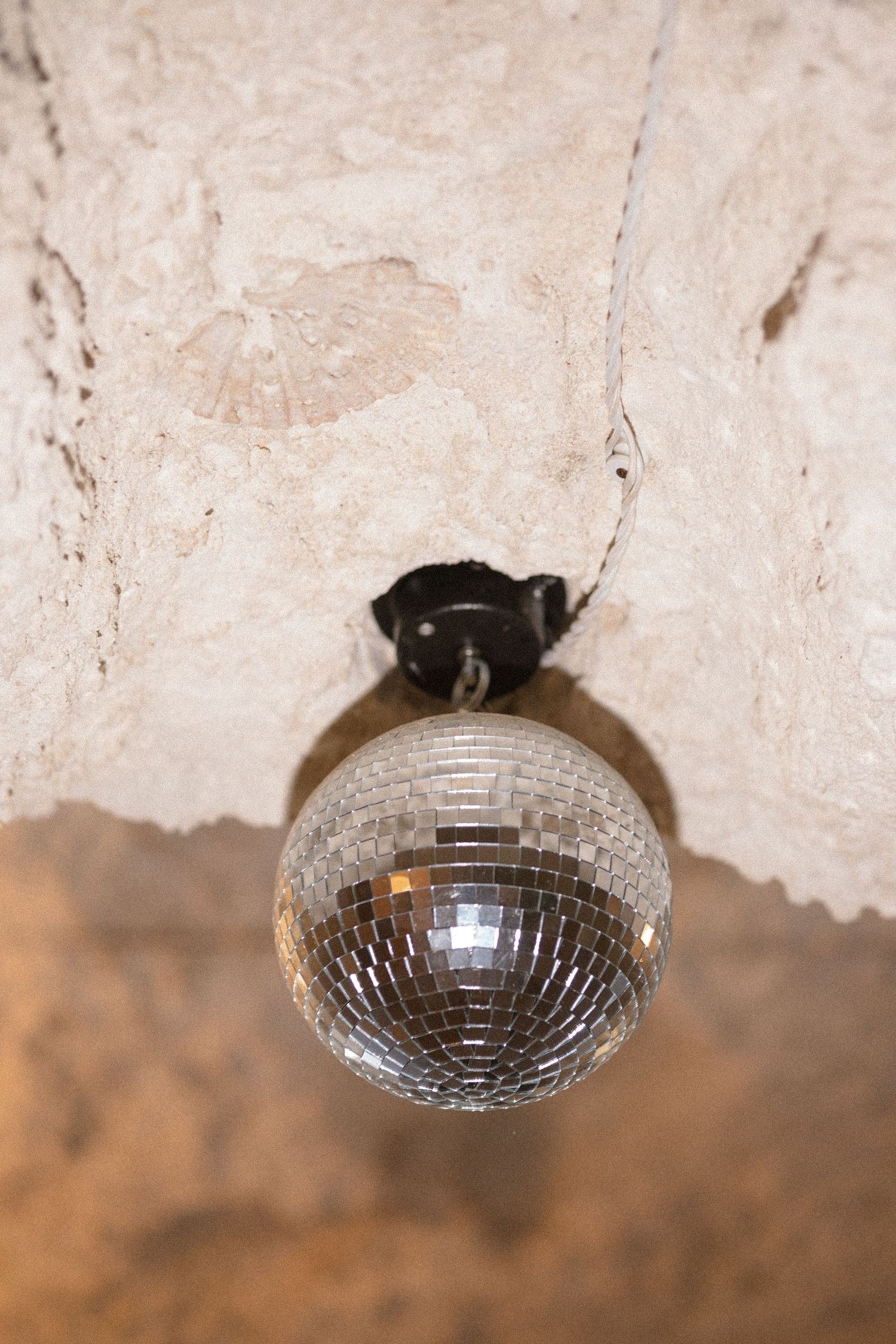 Silver mirror disco ball hanging from a black ceiling mount against a rough, textured whitewashed stone ceiling.
