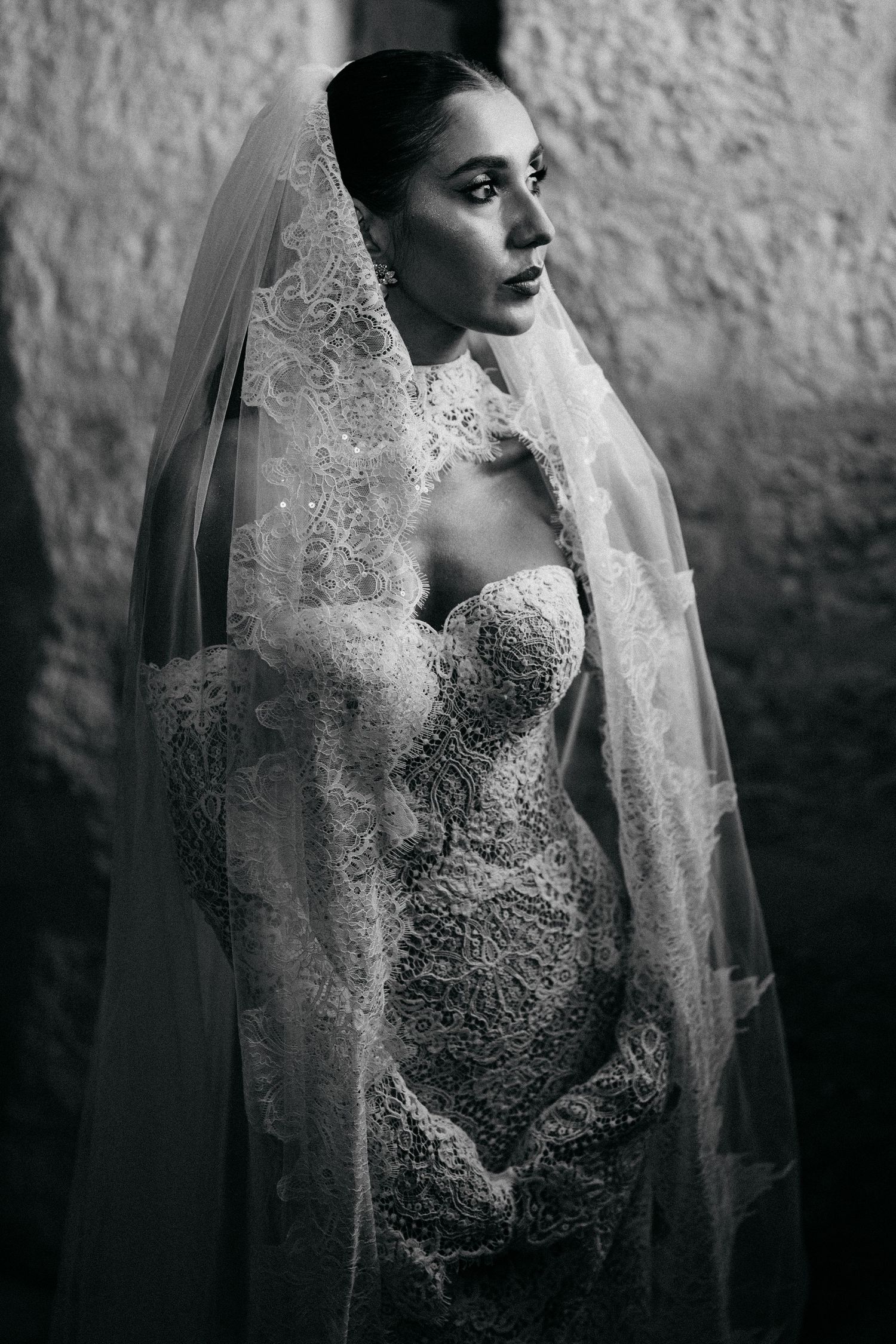 Black and white portrait of a bride in a lace gown and long veil gazing to the side against a rustic stone wall.