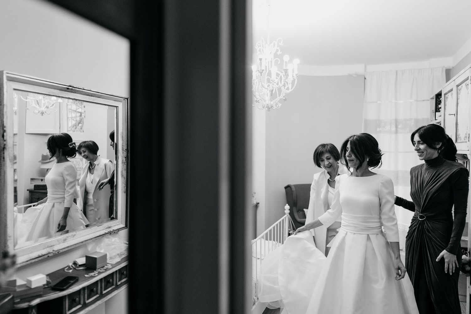 Black and white photo of a bride being helped into her wedding dress by bridesmaids in an elegant room.