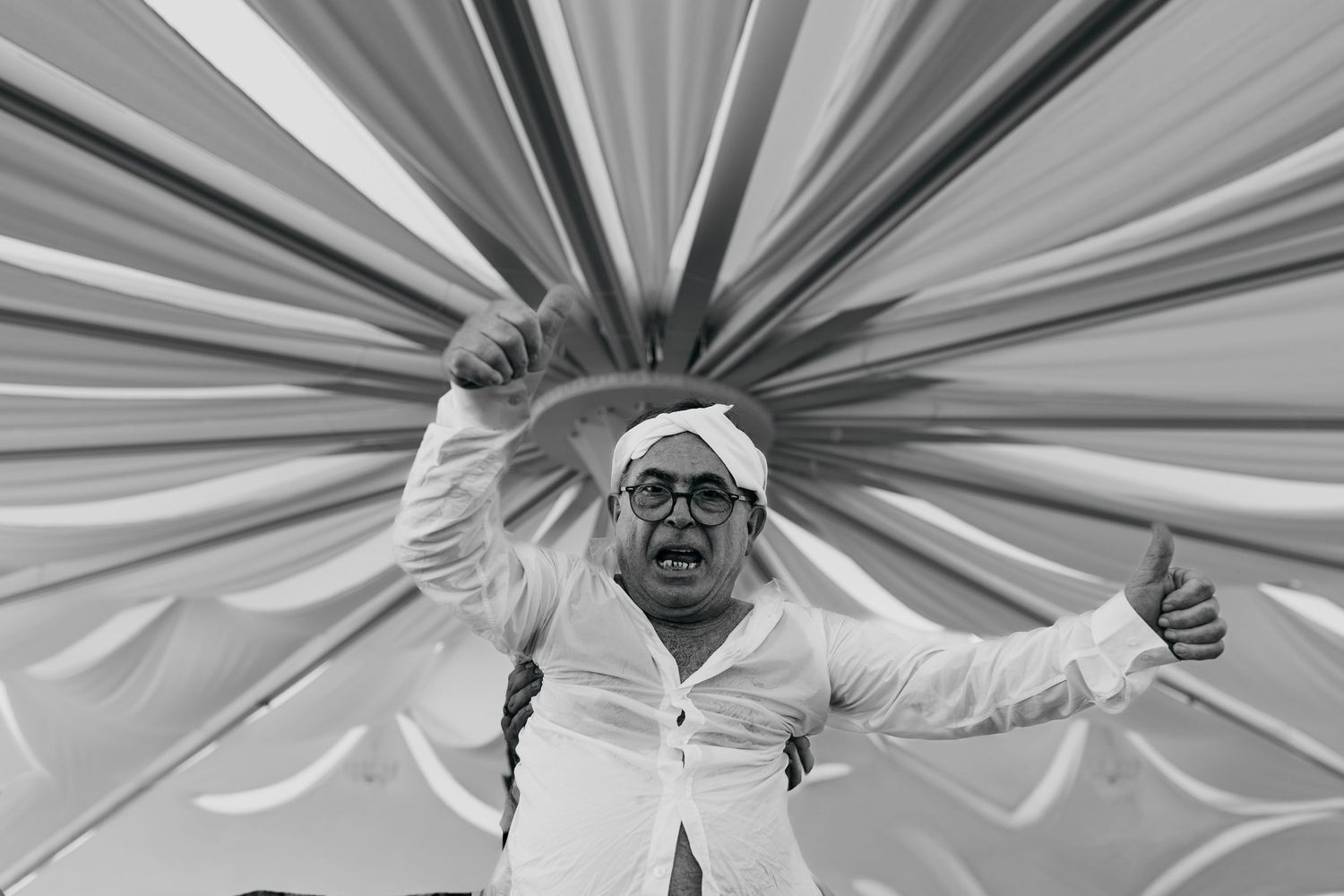 Black and white photo of a joyful man in white shirt and turban posing under colorful fabric streamers.