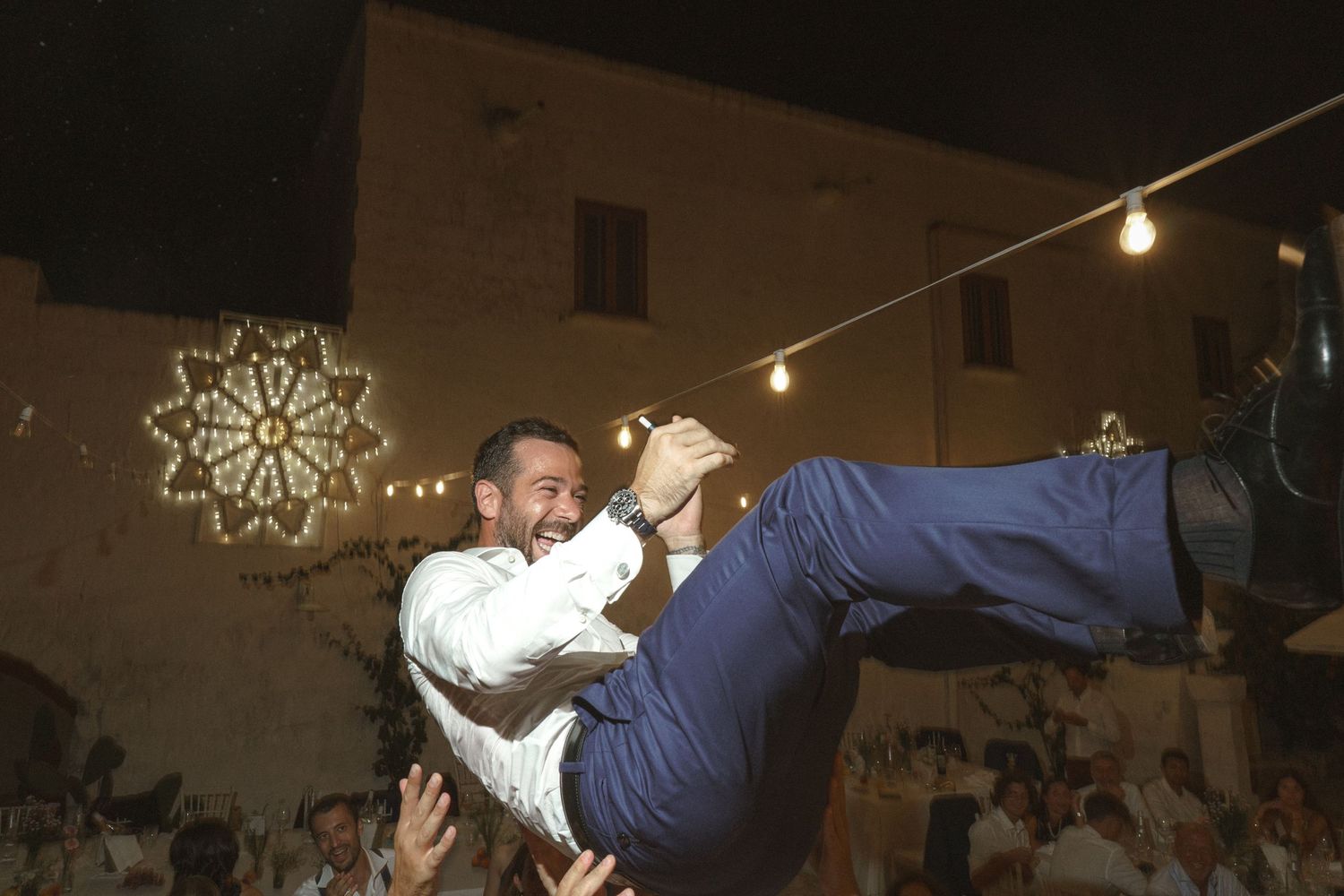Wedding guests lift and carry someone during an energetic nighttime celebration.