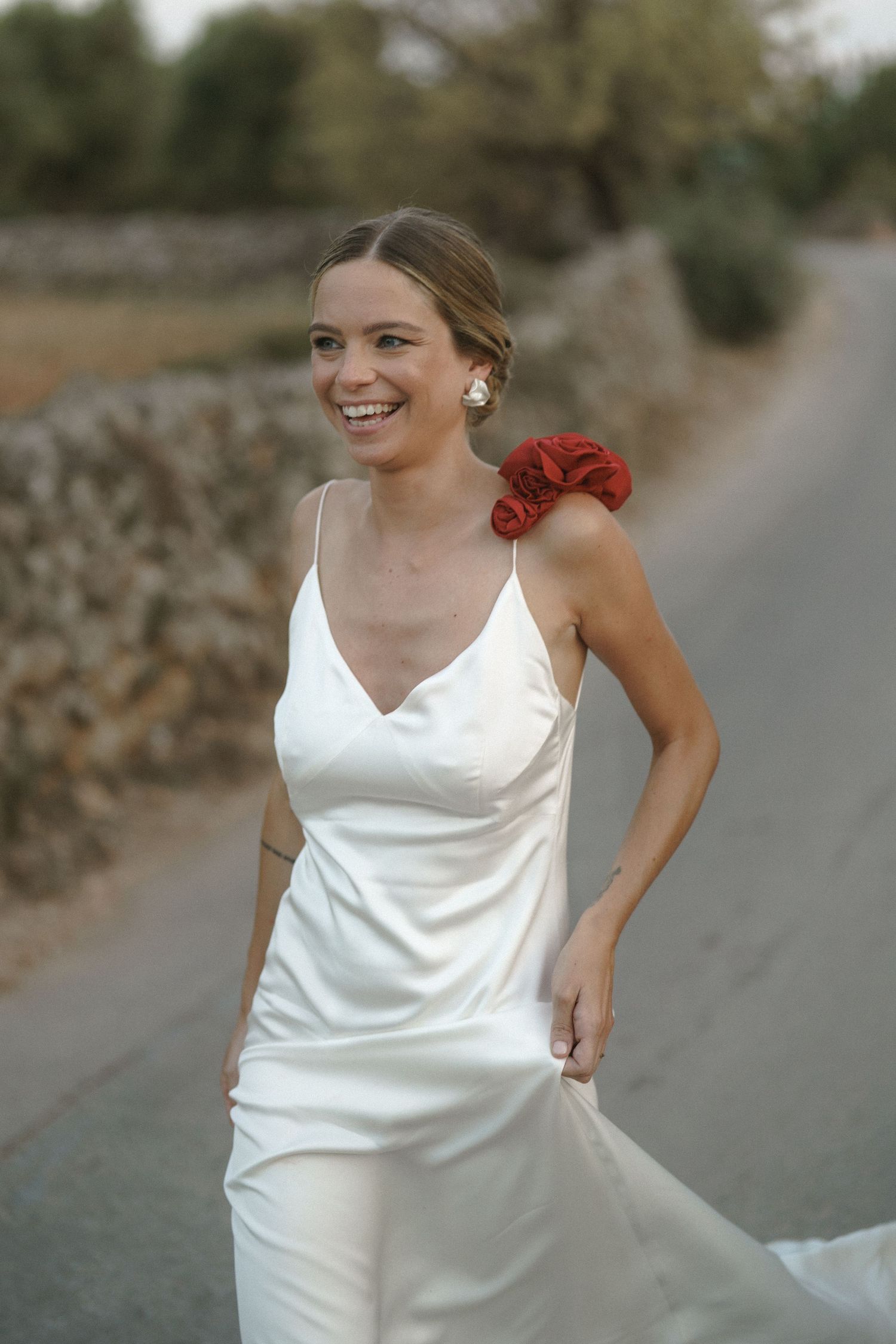 A white satin wedding dress with spaghetti straps and a red flower accent on the shoulder is worn on a country path.