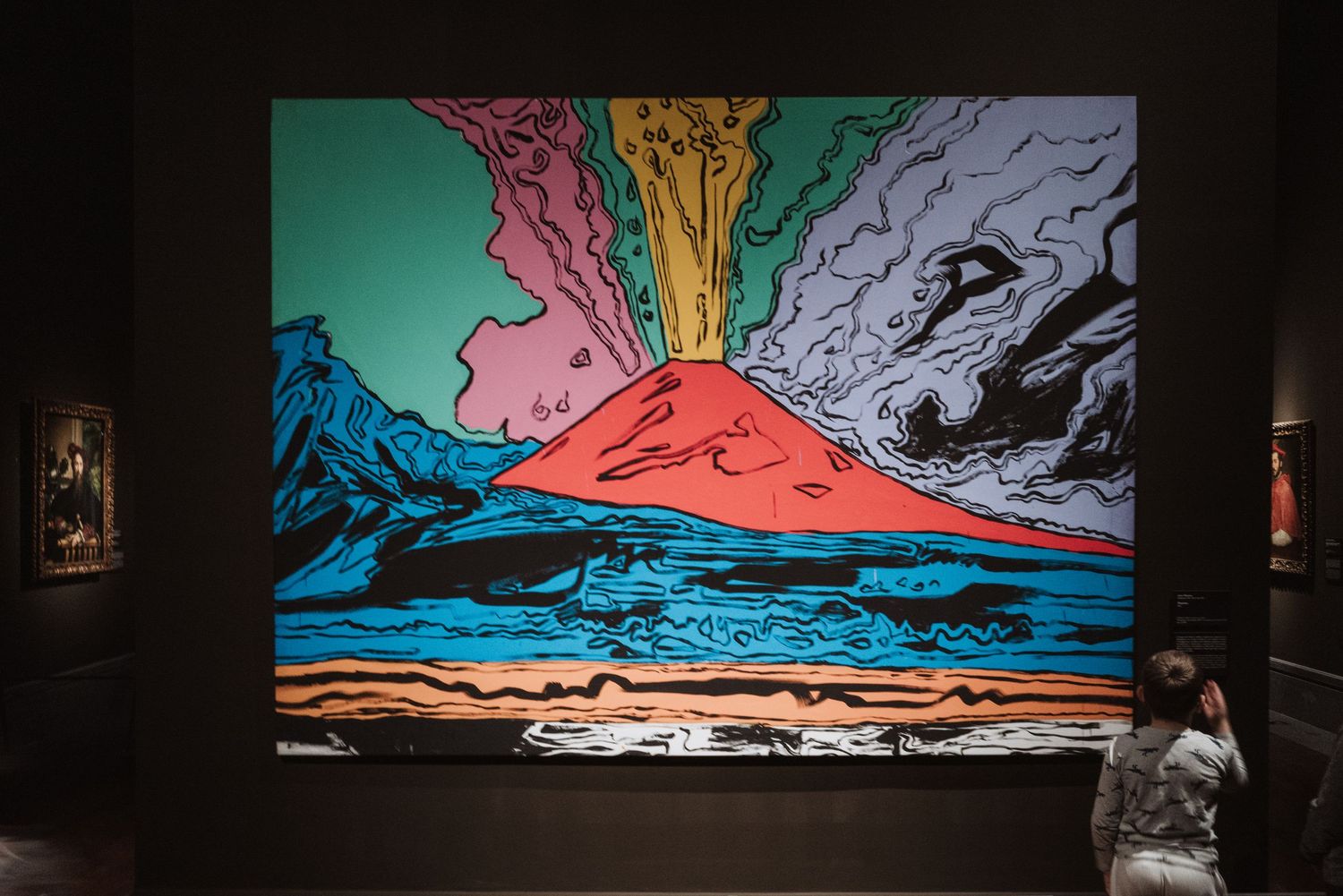 A person views a vibrant mural depicting an erupting volcano with colorful smoke plumes against a turquoise and blue landscape.