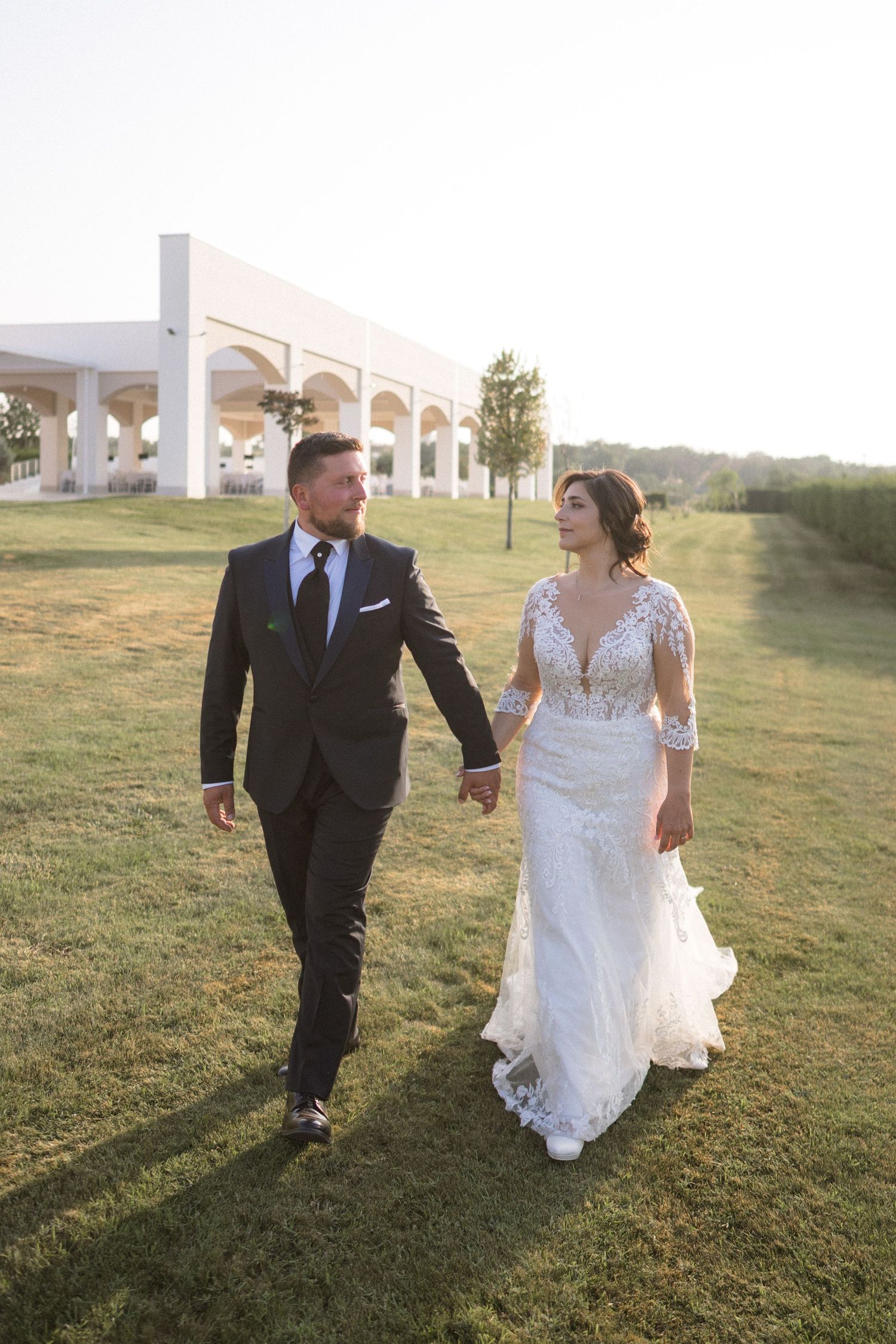 Newlyweds walking together on a grassy hill with white architectural columns in the background.