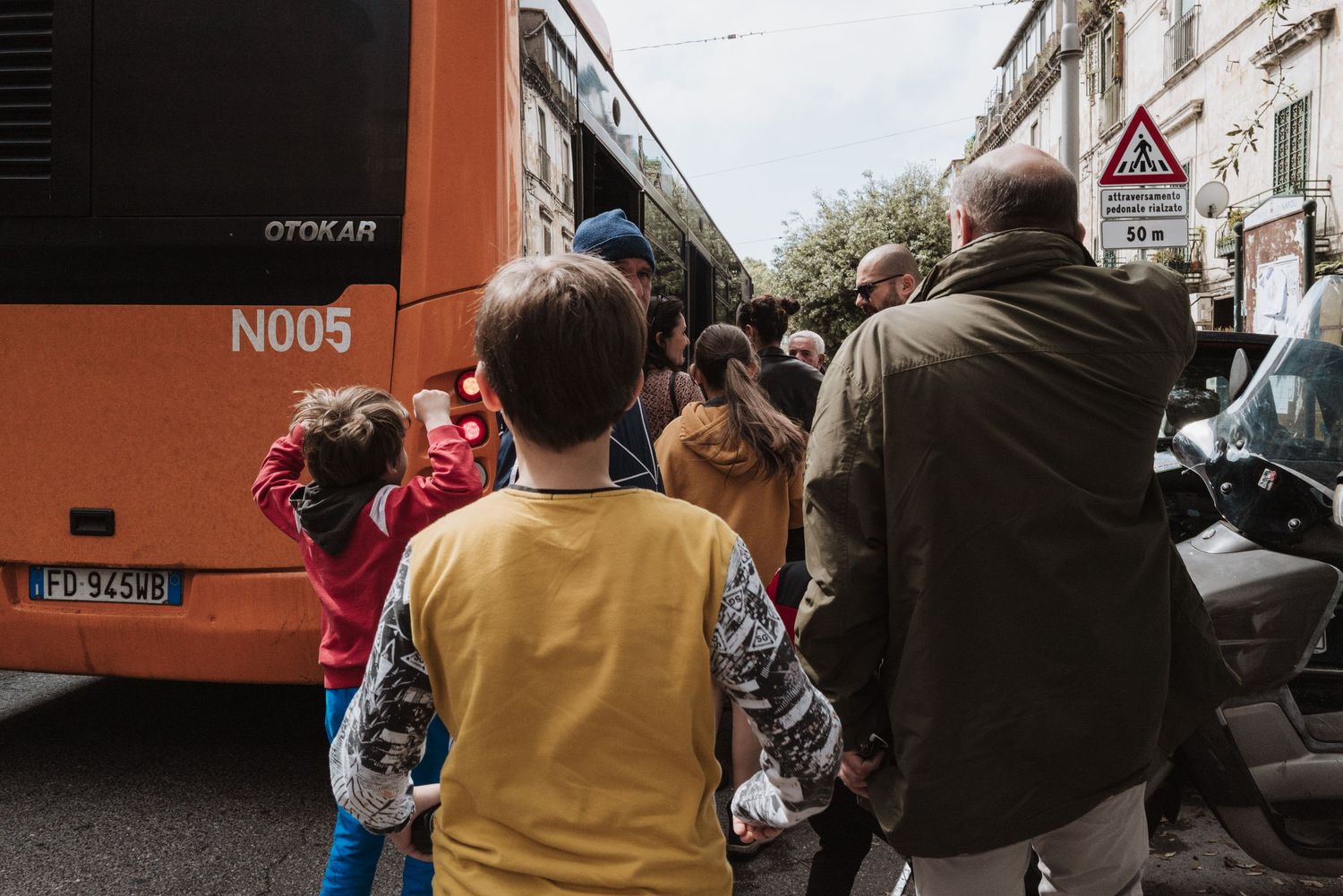 Group of people waiting to board an orange city bus on a busy street corner.