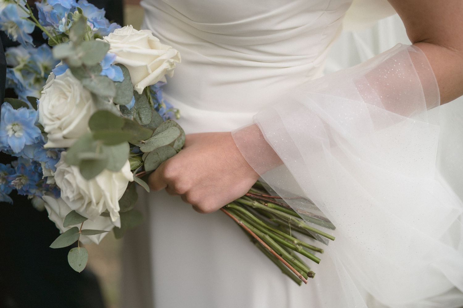 Close up of elegant white and blue wedding bouquet with roses and hydrangeas against bride's white dress.