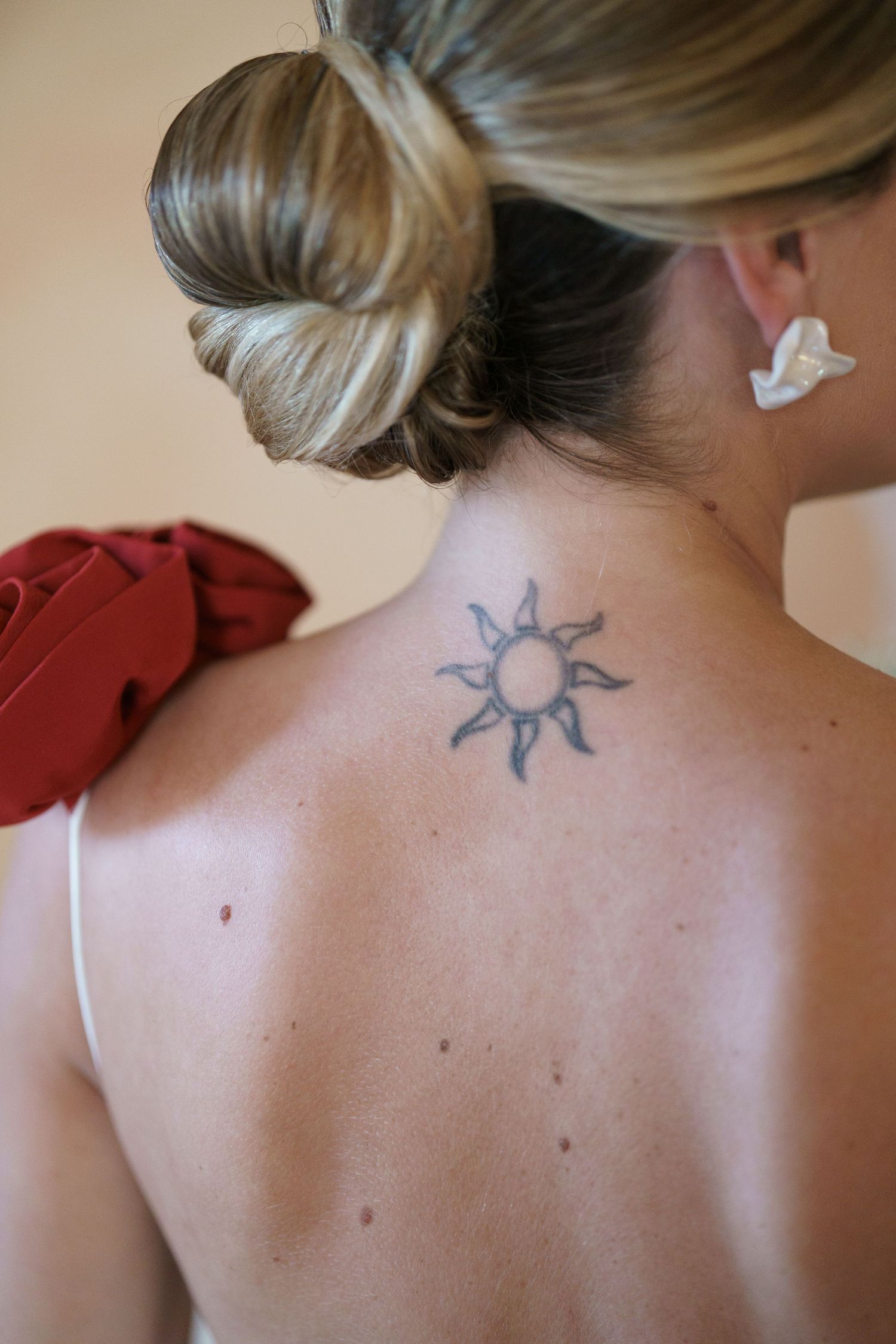 A sun tattoo design on the upper back with blonde hair in a bun and a dove-shaped earring visible.