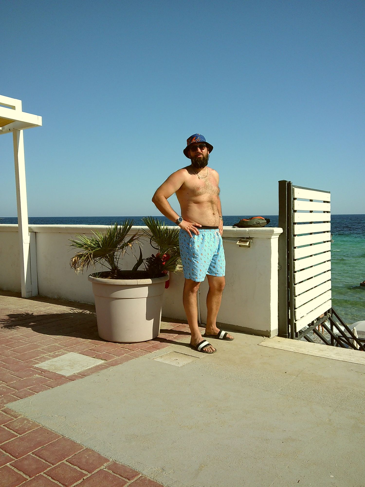 A person in swim shorts and hat standing on a beachfront terrace with ocean views.