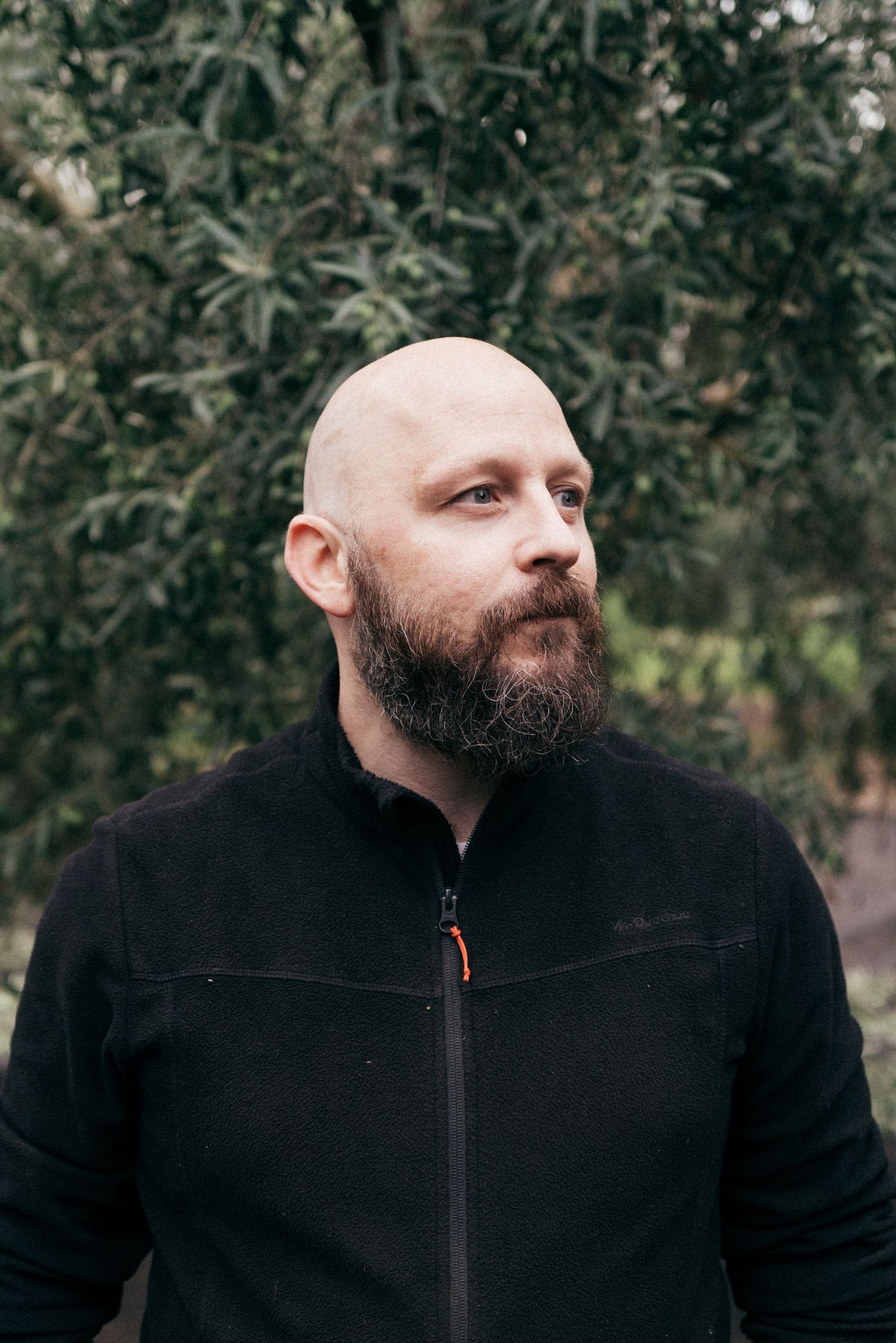 Portrait of a bald man with a dark beard wearing a black zip-up jacket against a natural outdoor background.