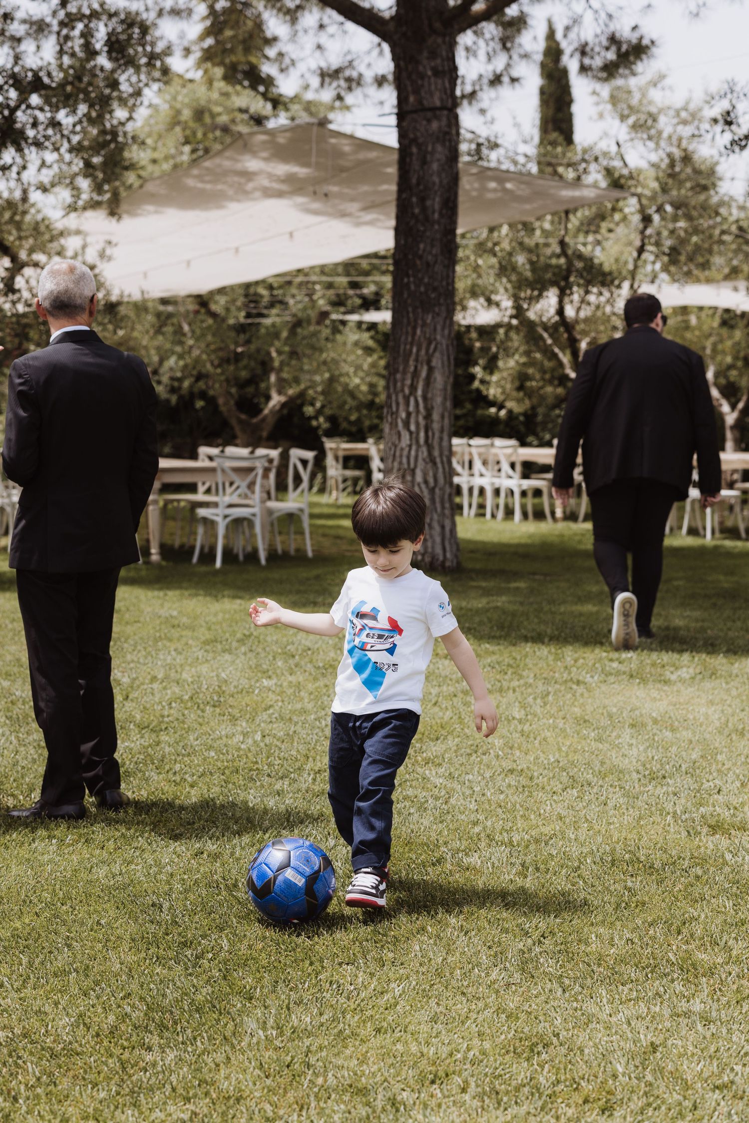 A young boy plays with a blue soccer ball on the lawn during a formal event.