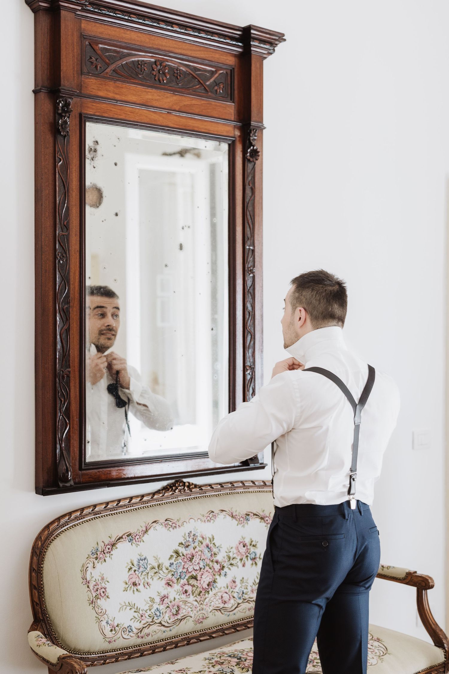 Person adjusting formal attire in ornate wooden framed mirror above vintage settee.