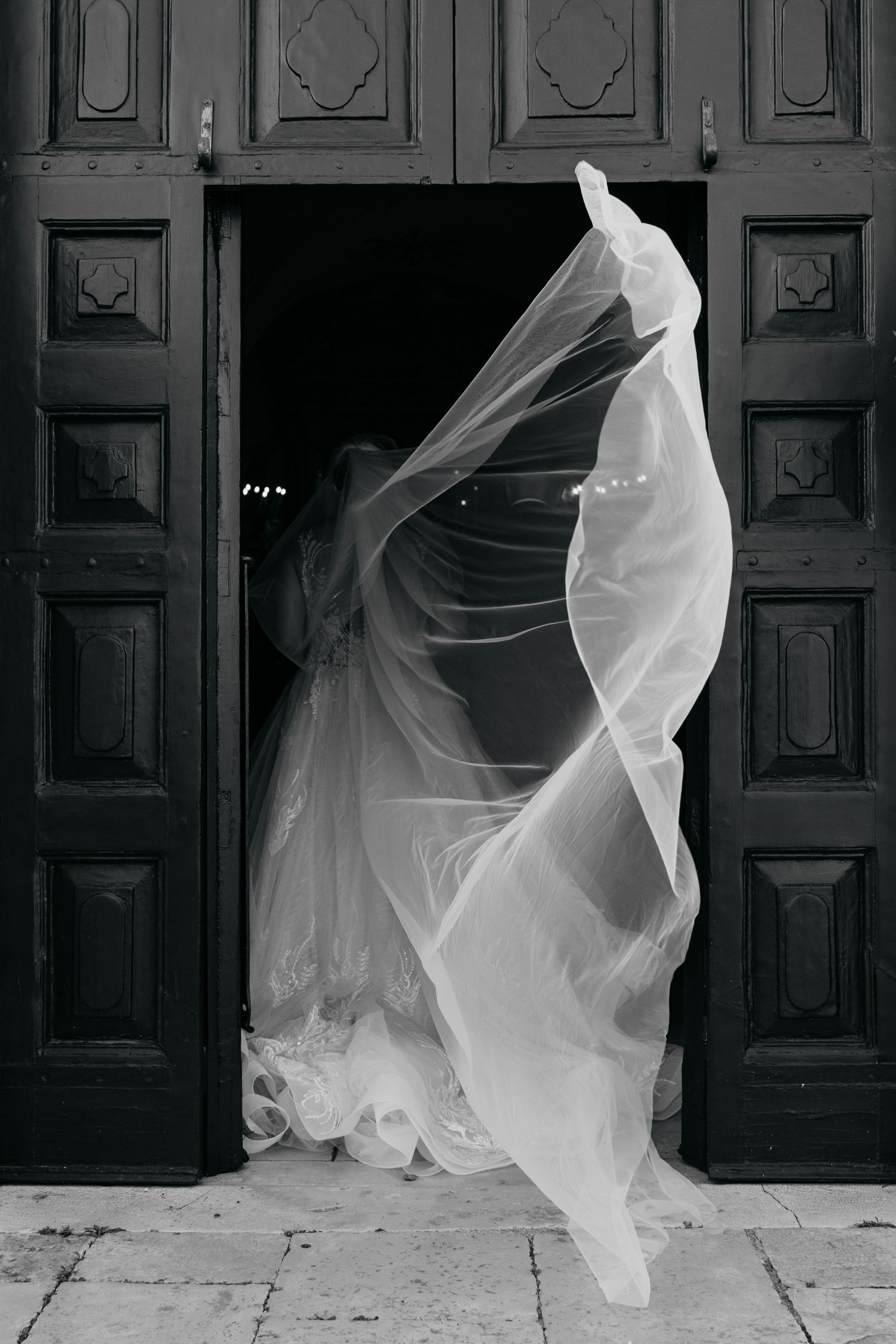 Flowing white wedding veil dramatically billowing against dark wooden doors.