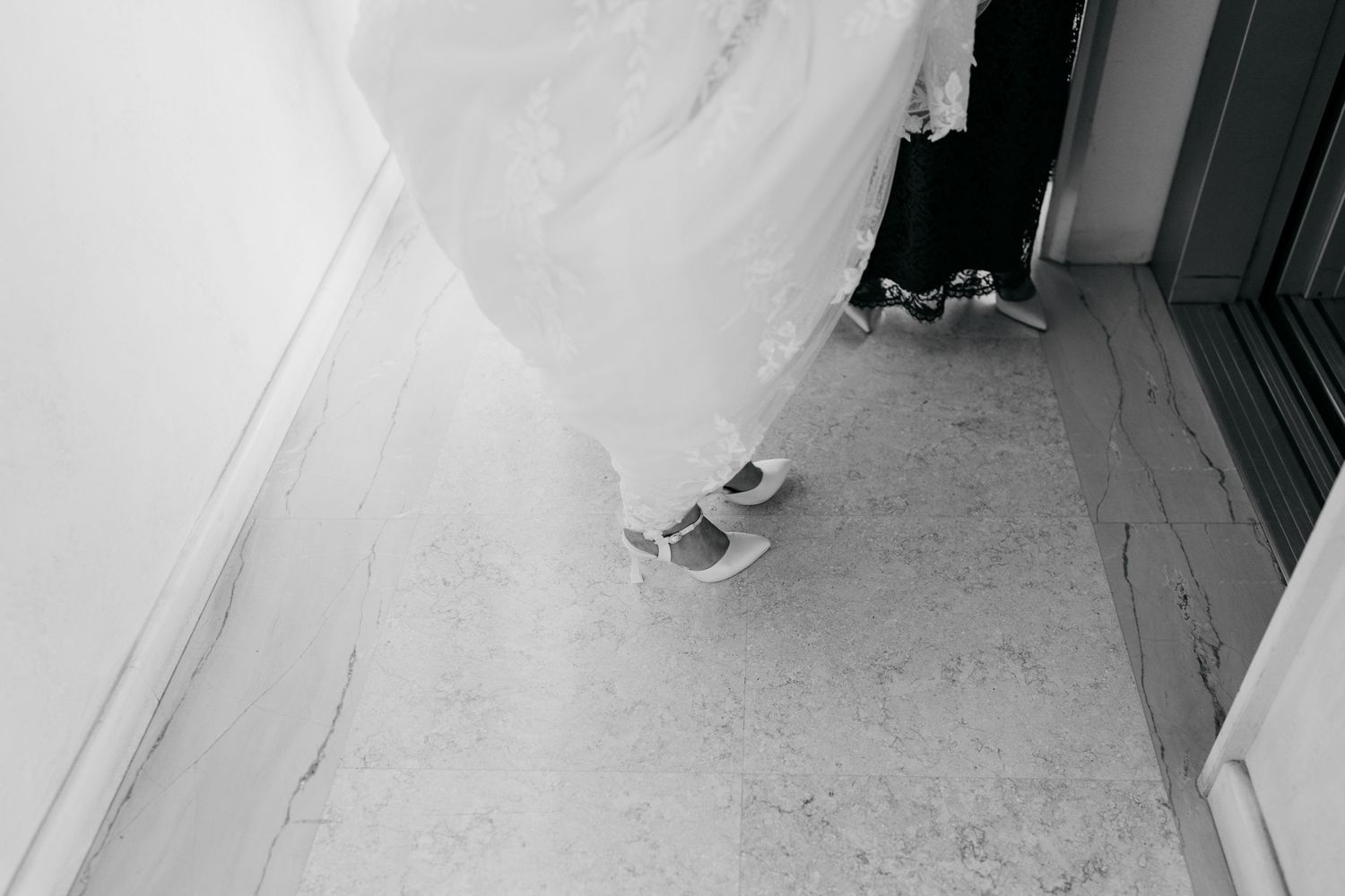 Black and white photo of a bride in a white wedding gown wearing casual sneakers, standing on a tiled floor.