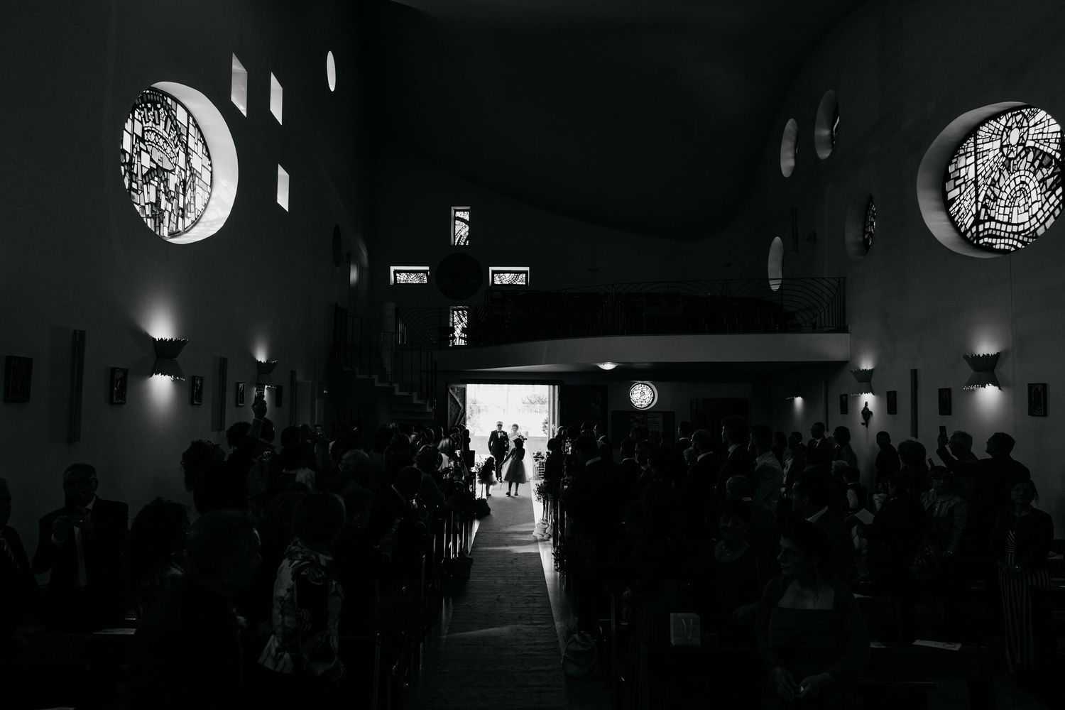 Black and white photo of a church wedding ceremony with guests seated, stained glass windows, and bright altar light.
