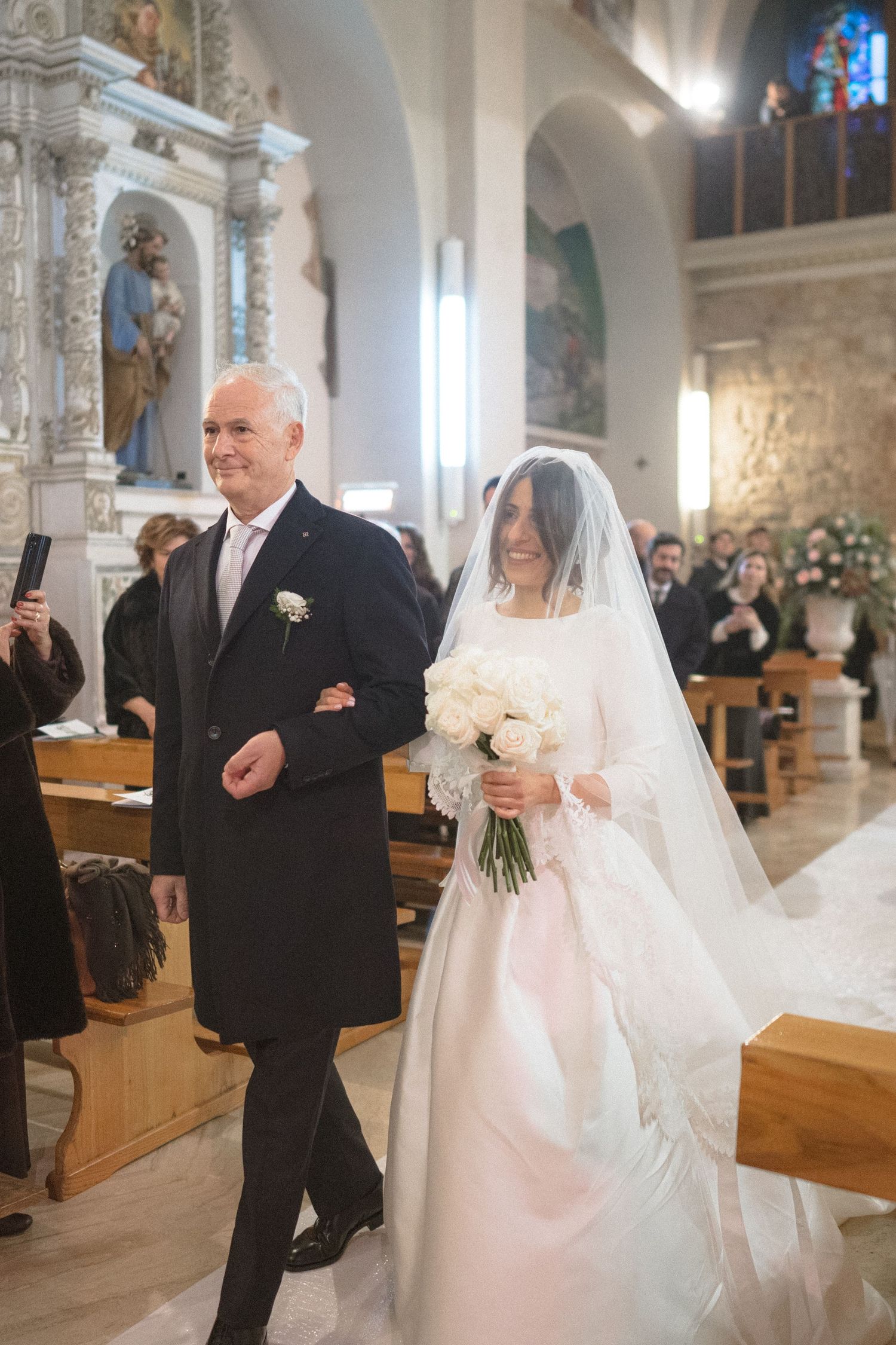 Smiling bride in white gown and veil walks down church aisle with father during wedding ceremony.