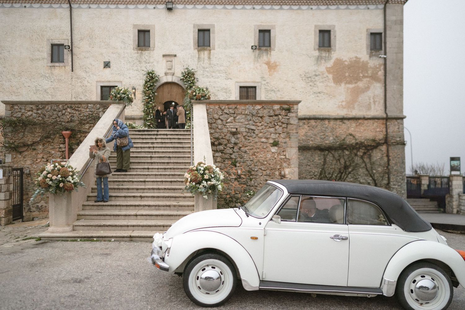 White vintage Volkswagen Beetle parked outside a rustic stone wedding venue with floral decor.