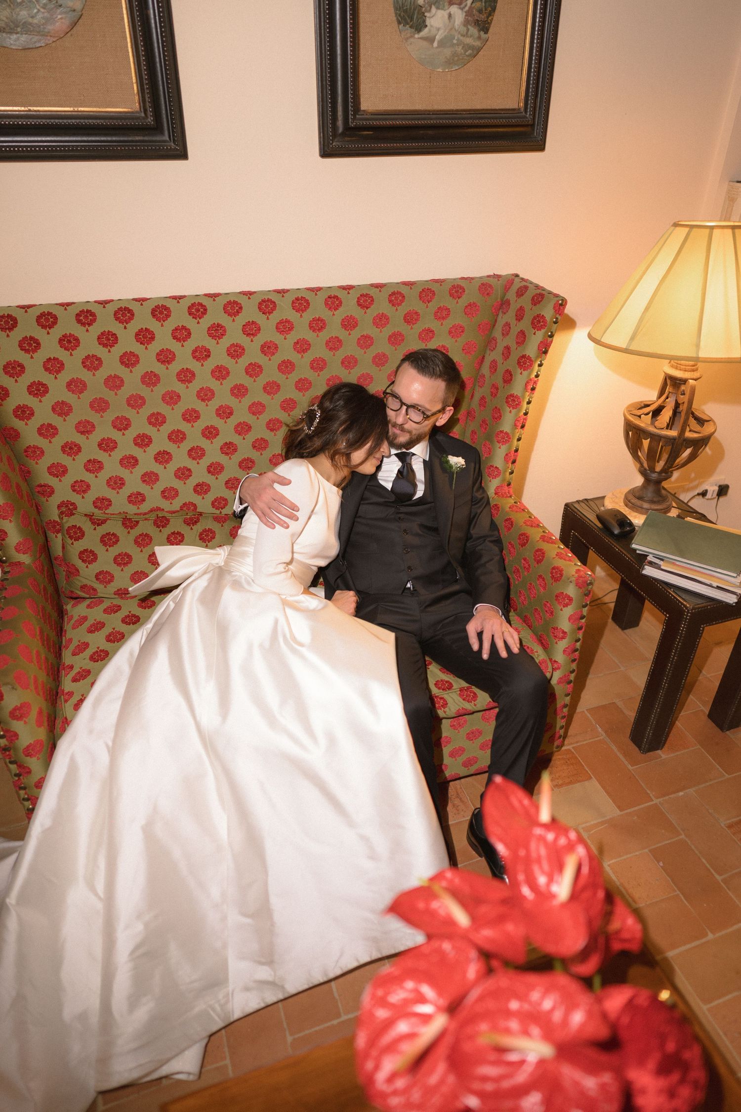 Bride and groom sitting closely together on an ornate red sofa in an elegantly decorated room.
