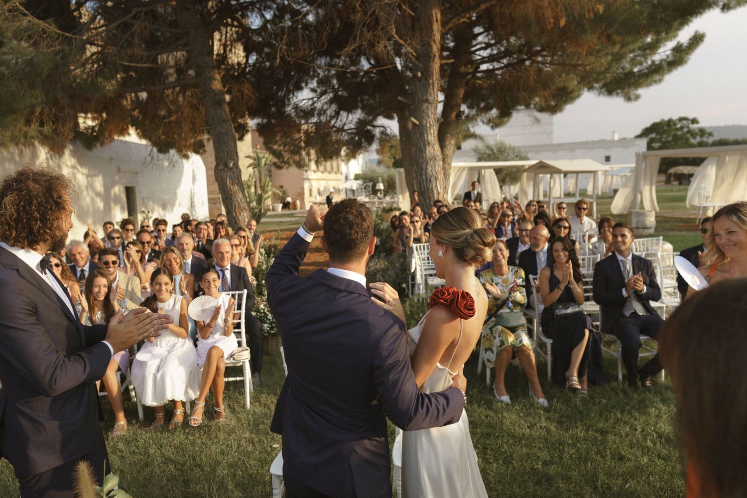 A crowd gathers outdoors under tall pine trees during an evening wedding ceremony with string lights overhead.