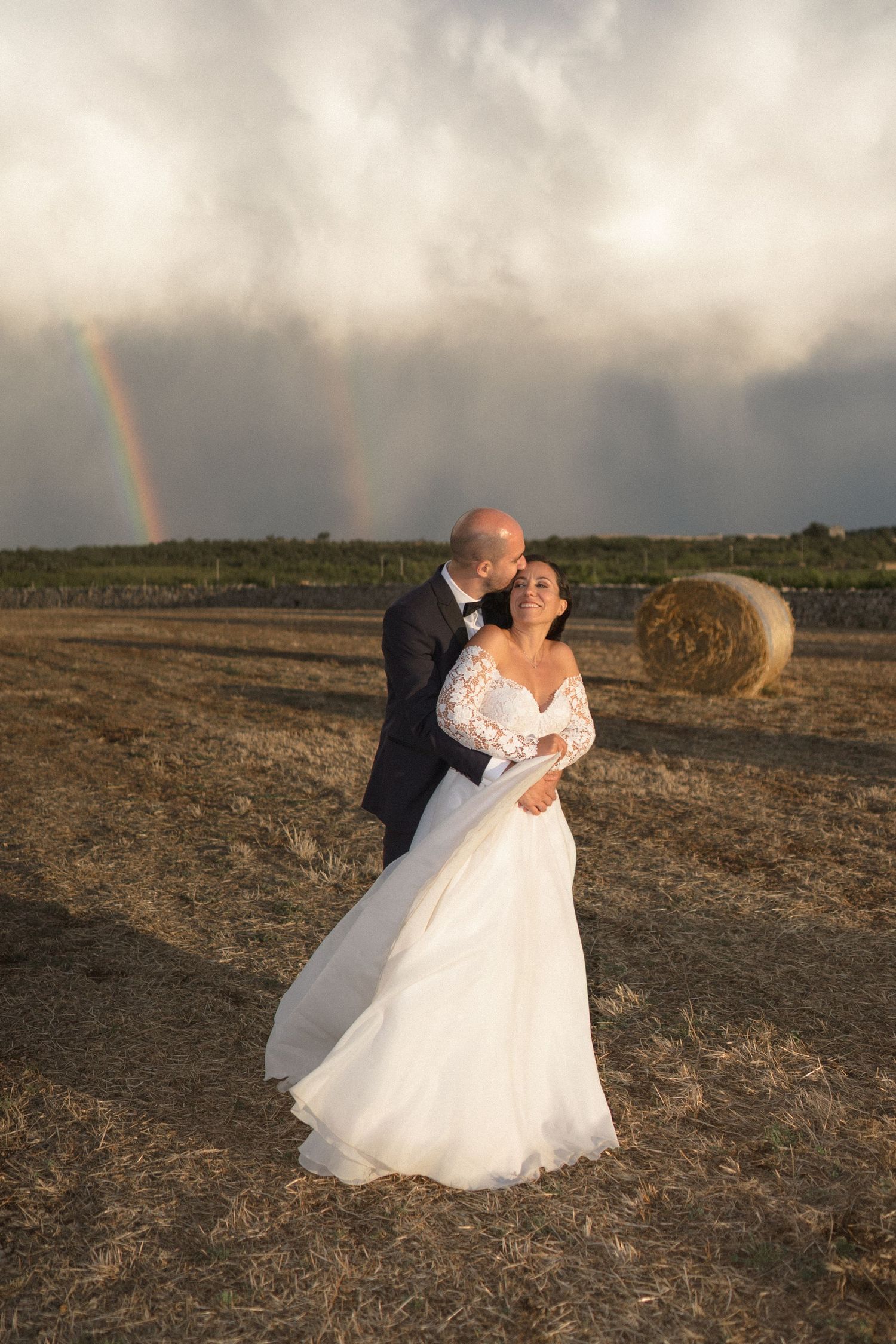 A couple in wedding attire shares a romantic kiss in a field at sunset with a rainbow visible in the background.