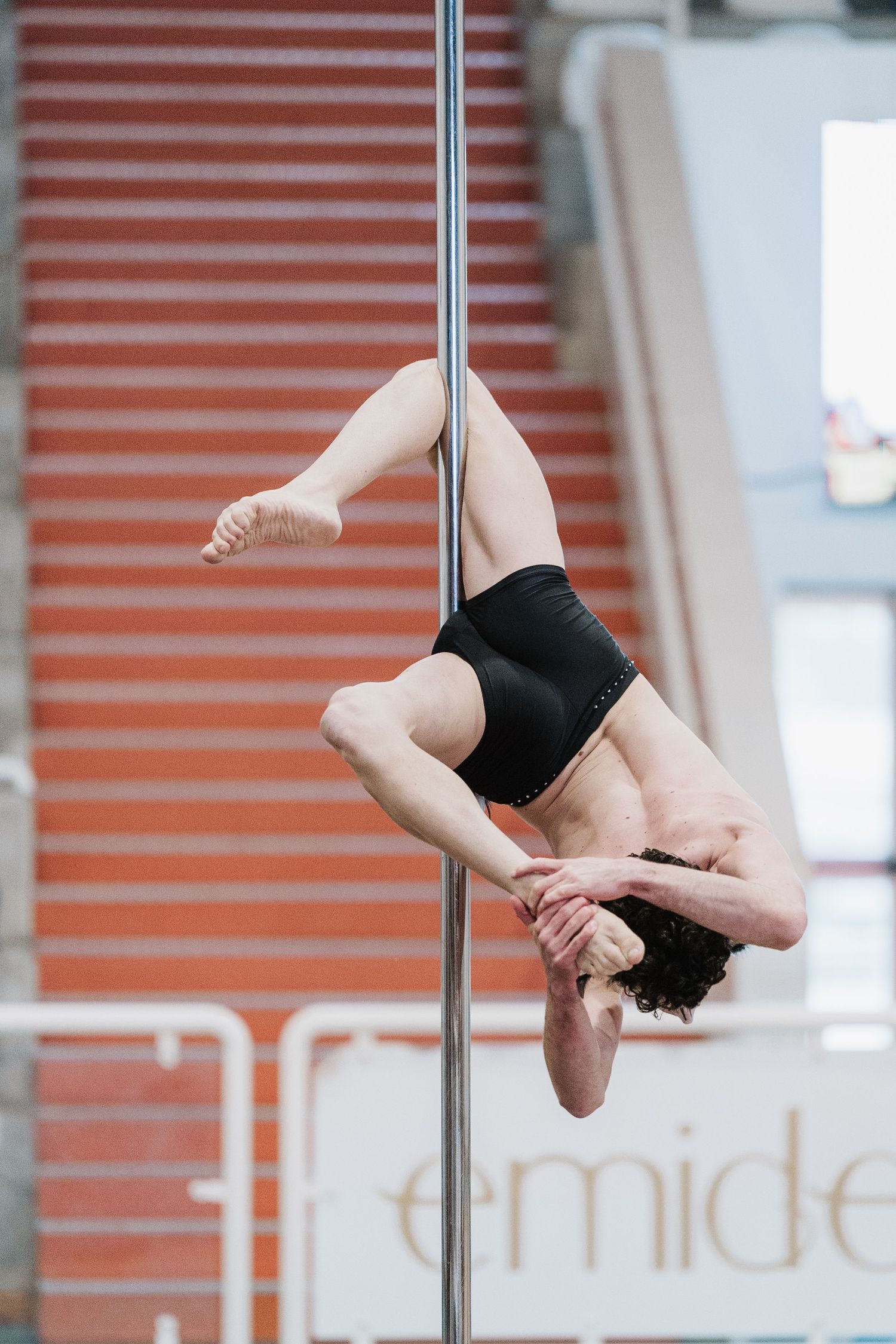 Athletic performer in black attire demonstrates strength and grace during a pole dance routine against an orange wall.