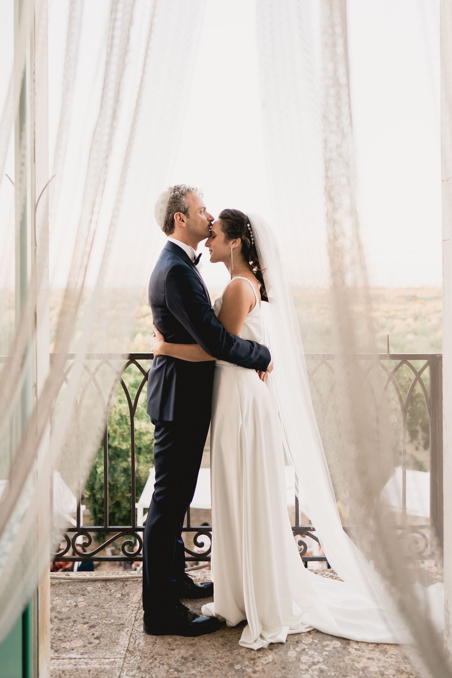 A romantic wedding couple embraces by sheer white curtains on a balcony in an elegant setting.