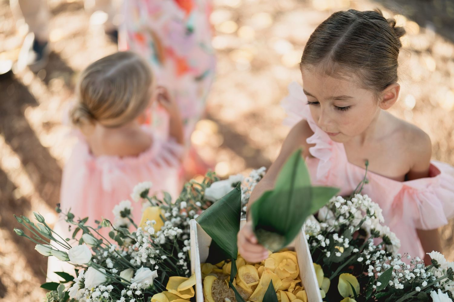 Young attendants in pink off-shoulder dresses examine delicate white and green floral bouquets at an outdoor event.