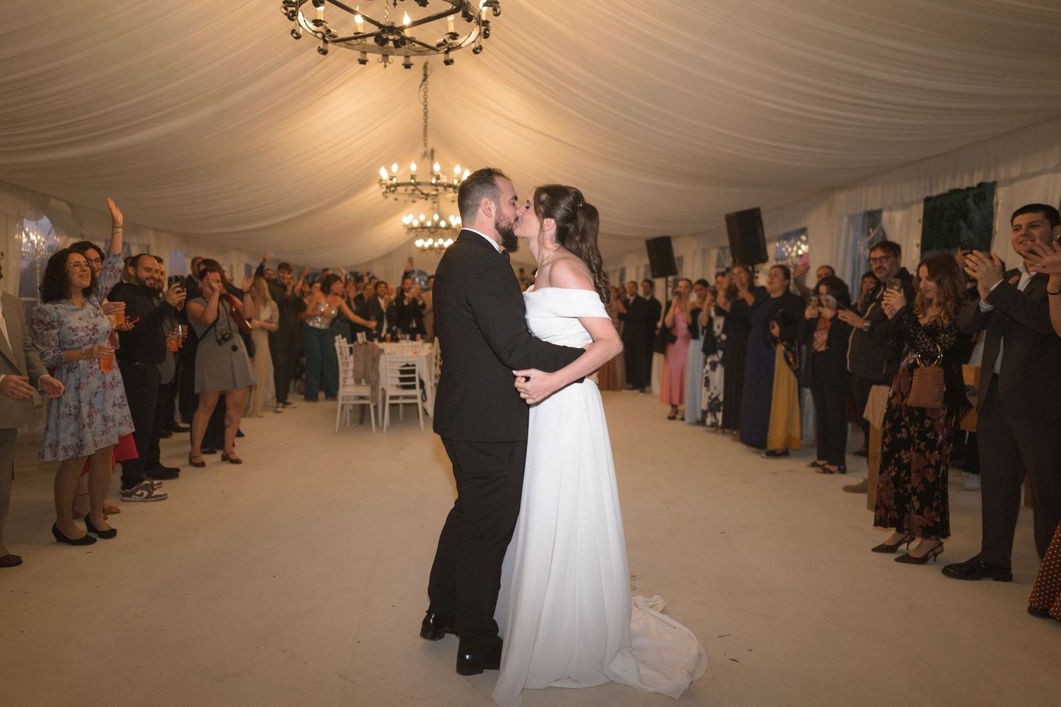 Wedding guests form a circle around a couple dancing in an elegantly draped reception tent with chandeliers.