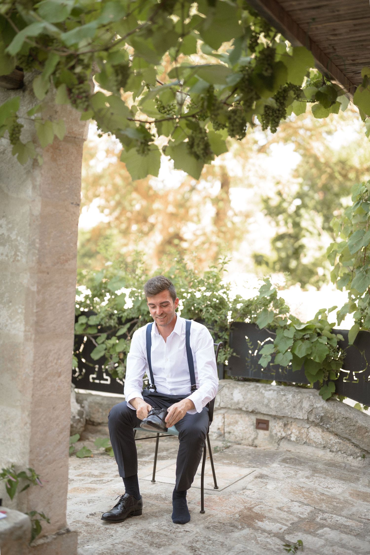 Person in formal attire with suspenders sitting on a stone bench under green foliage.