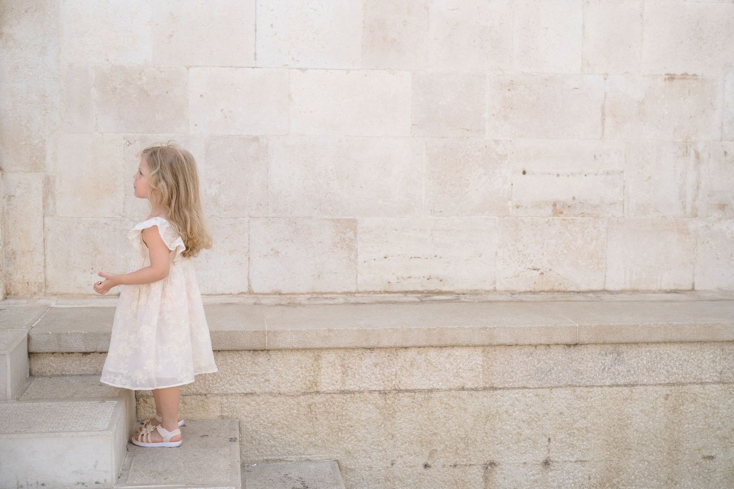 A small child in a white dress walks down a stone corridor with light filtering in from above.