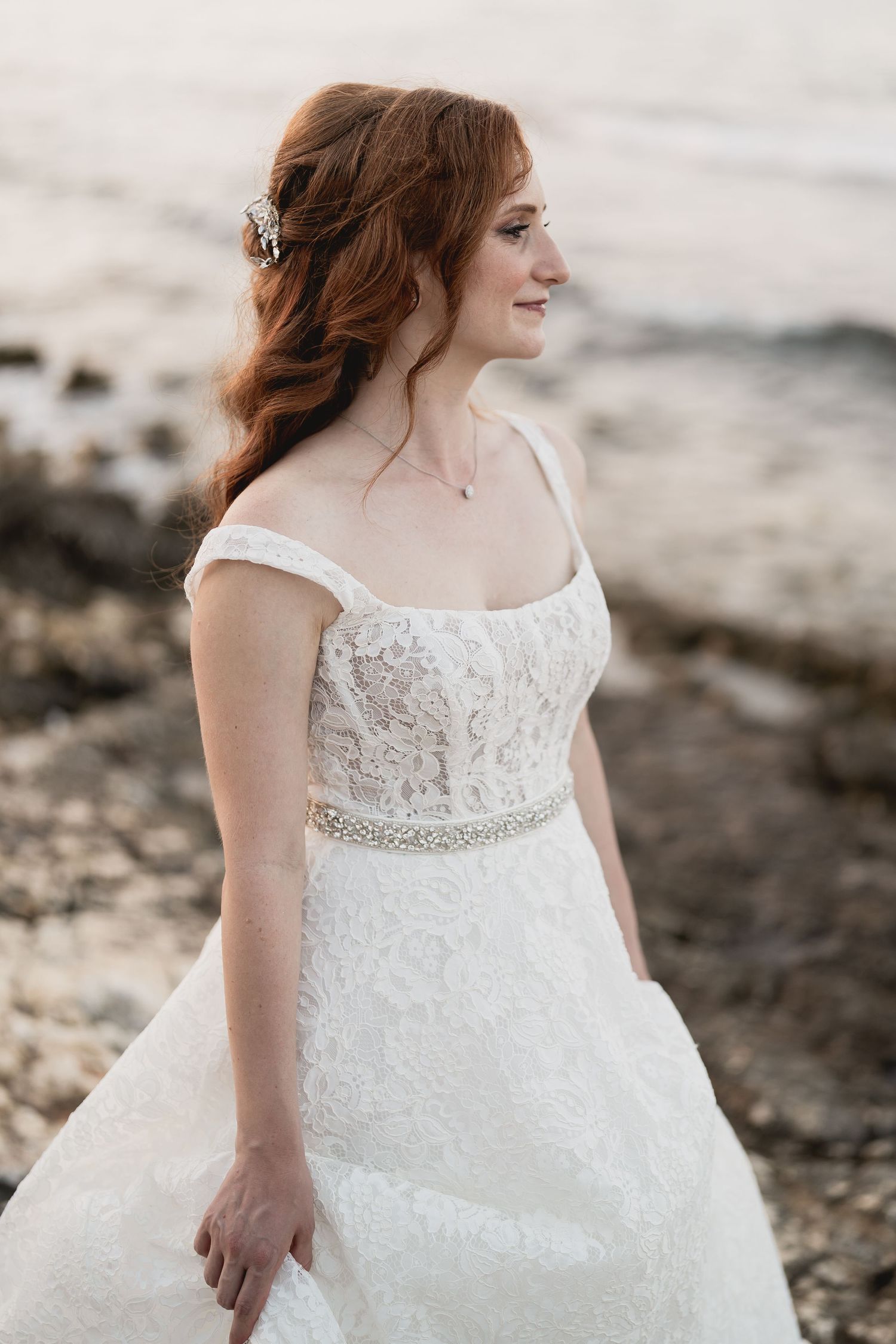 Bride in a white lace wedding dress with beaded belt stands by the ocean.