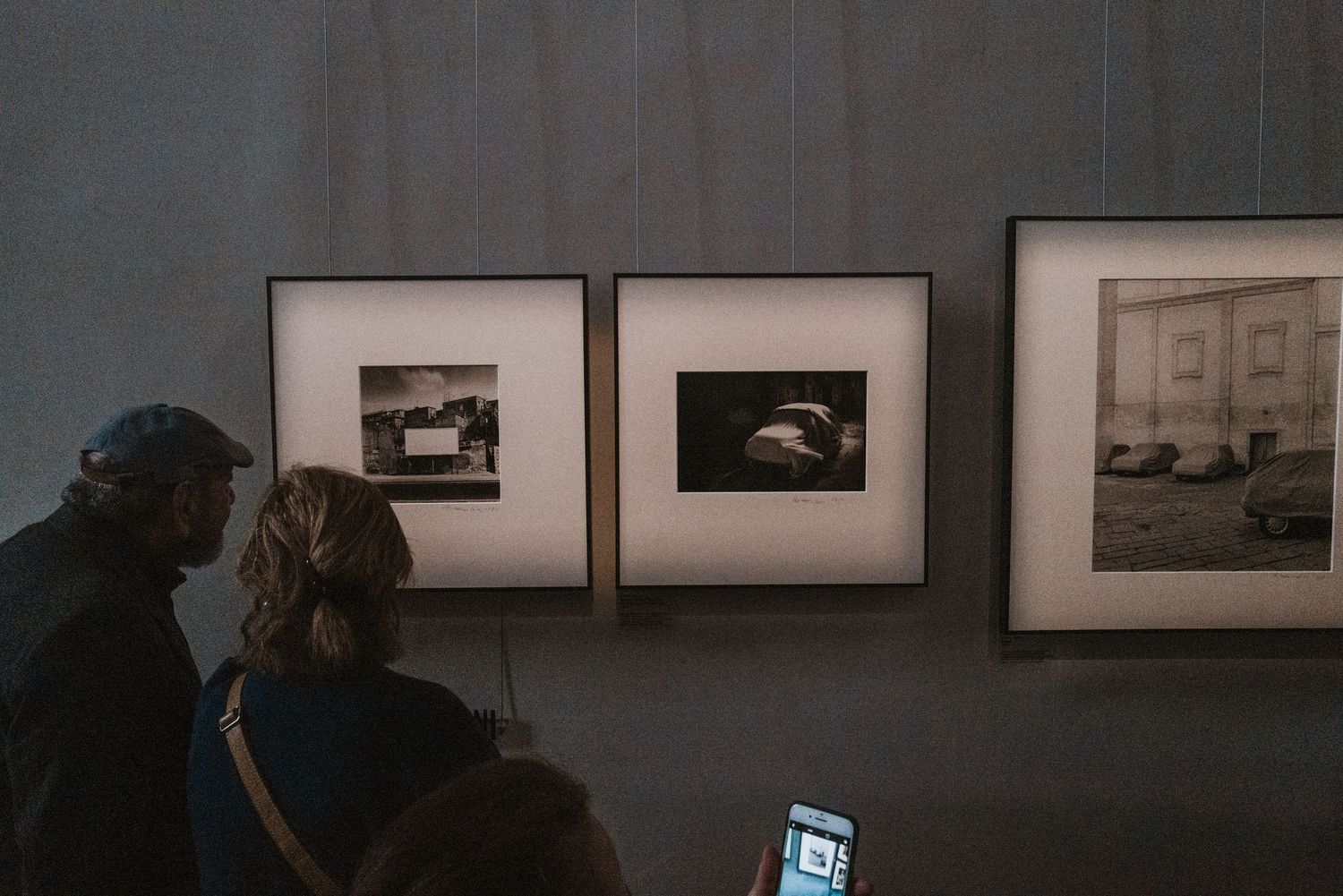 Gallery visitors view black and white photographs displayed on a dark wall in a dimly lit exhibition space.