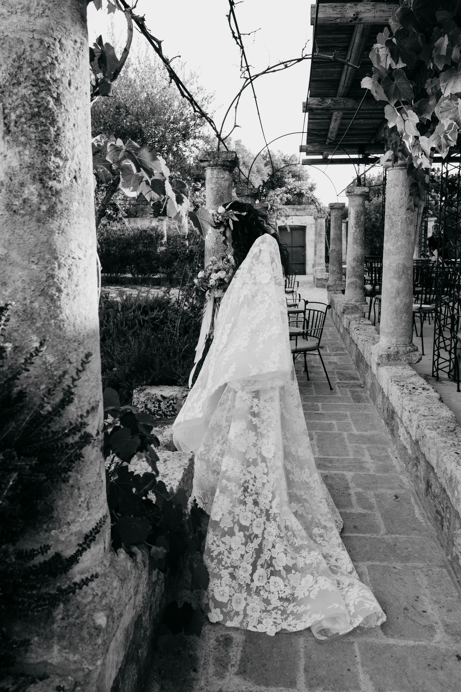 A flowing lace wedding dress captured in black and white against stone pillars and tiled patio.