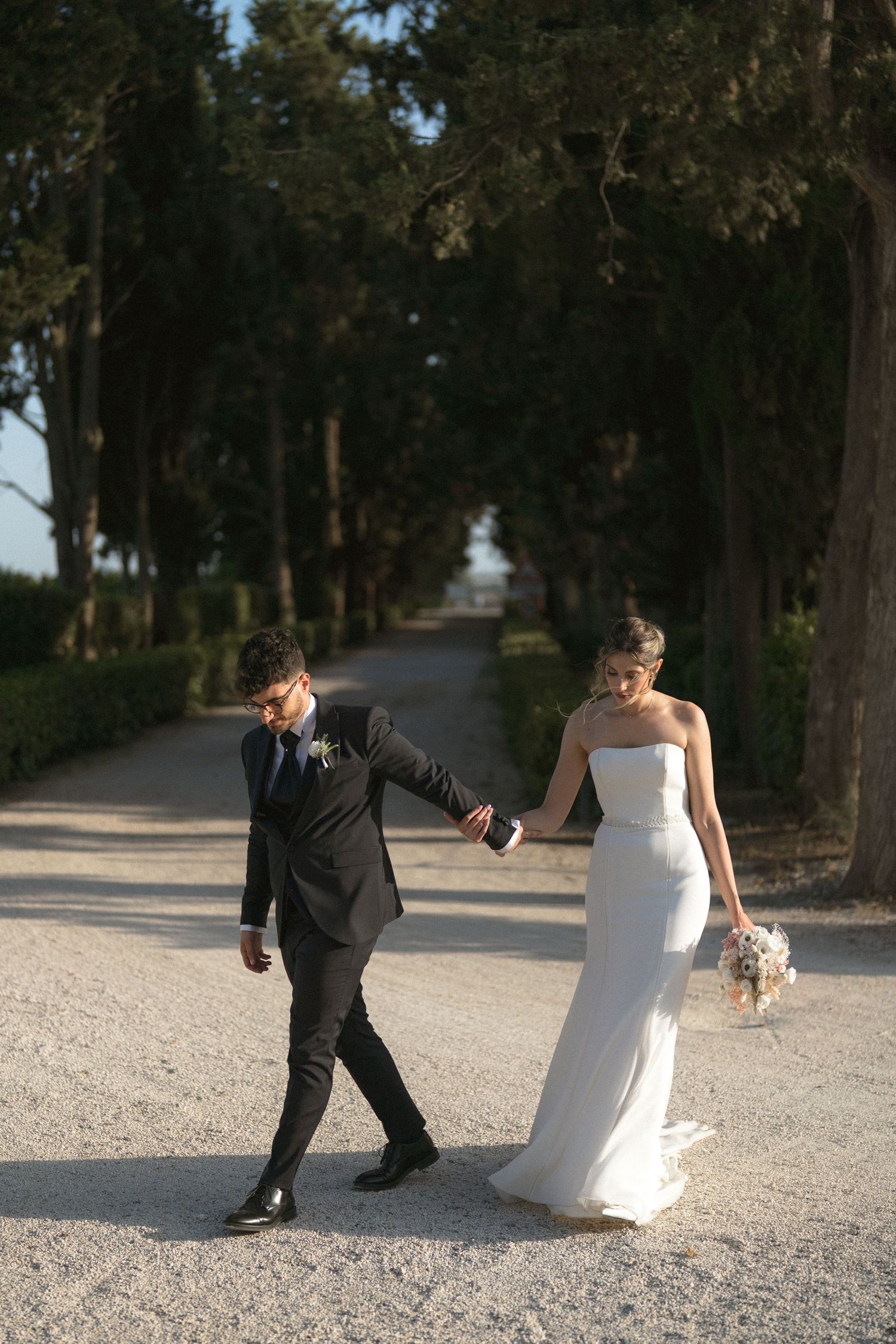 Playful wedding couple walking and dancing on a tree-lined gravel path during their photo session.