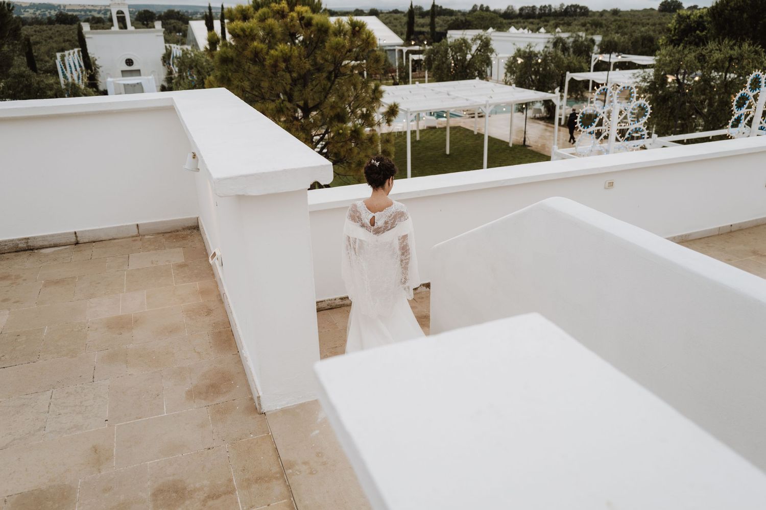 A figure in a white dress stands on a rooftop terrace with white architectural details and greenery in the background.