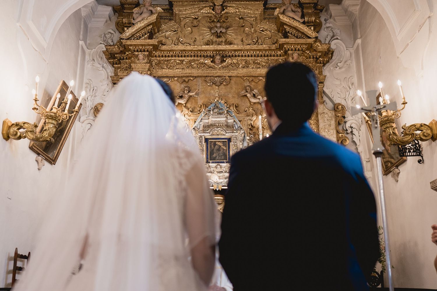 A wedding ceremony view from behind in an ornate church with gilded altar and wall sconces.