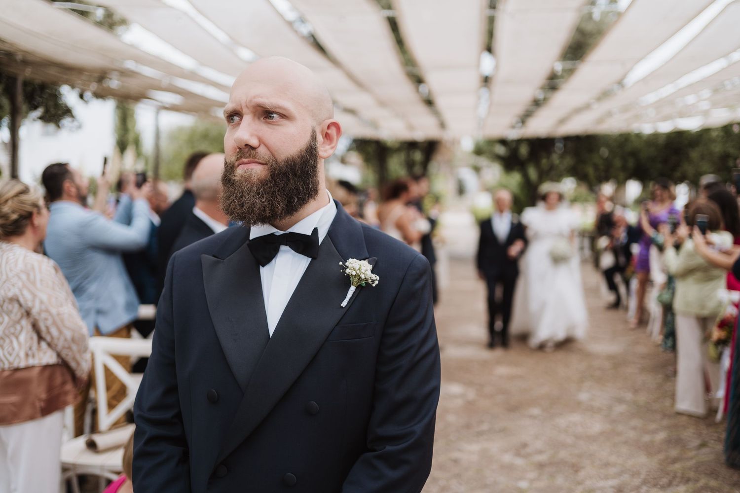 A groom in black tuxedo with boutonniere at outdoor wedding reception.