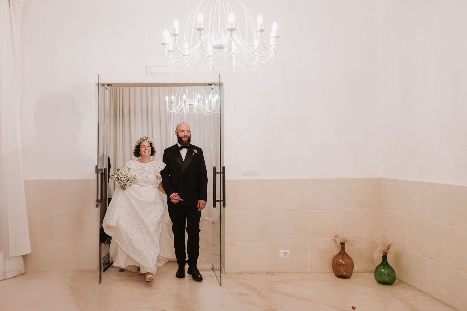 A couple makes their grand entrance through double doors in an elegant white reception room.