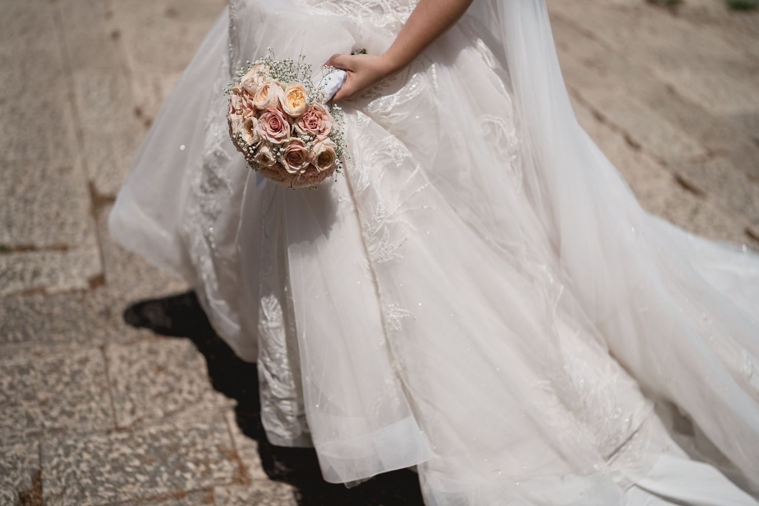 Close-up of an elegant white wedding dress with a blush-colored floral bouquet.