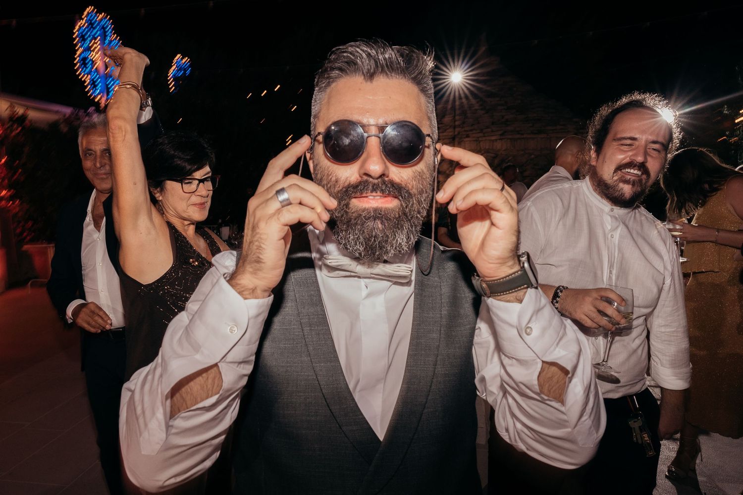 Stylish party guest in sunglasses and formal attire adjusts headphones during celebration.