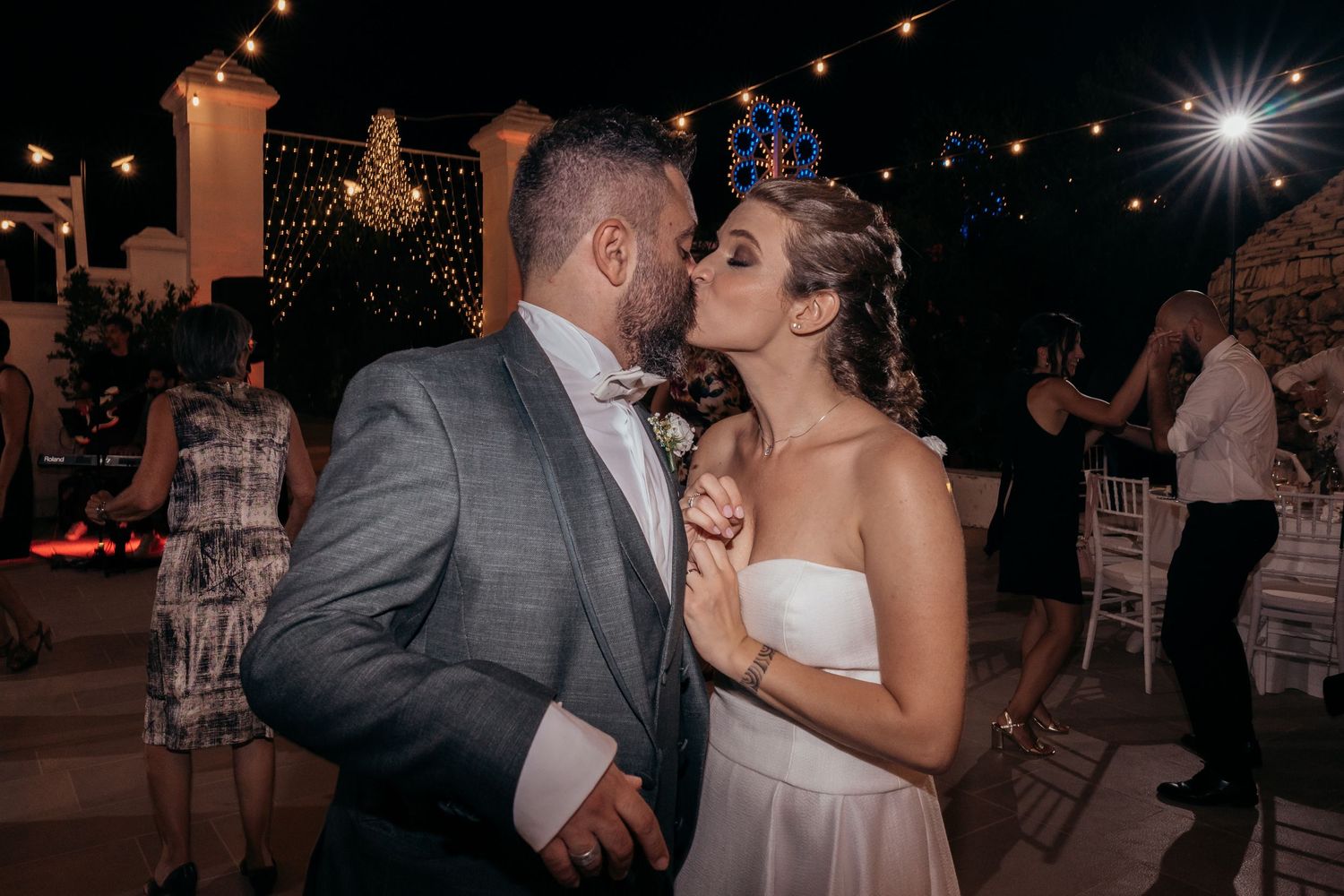 A couple shares a romantic moment while dancing under string lights at their outdoor wedding reception.