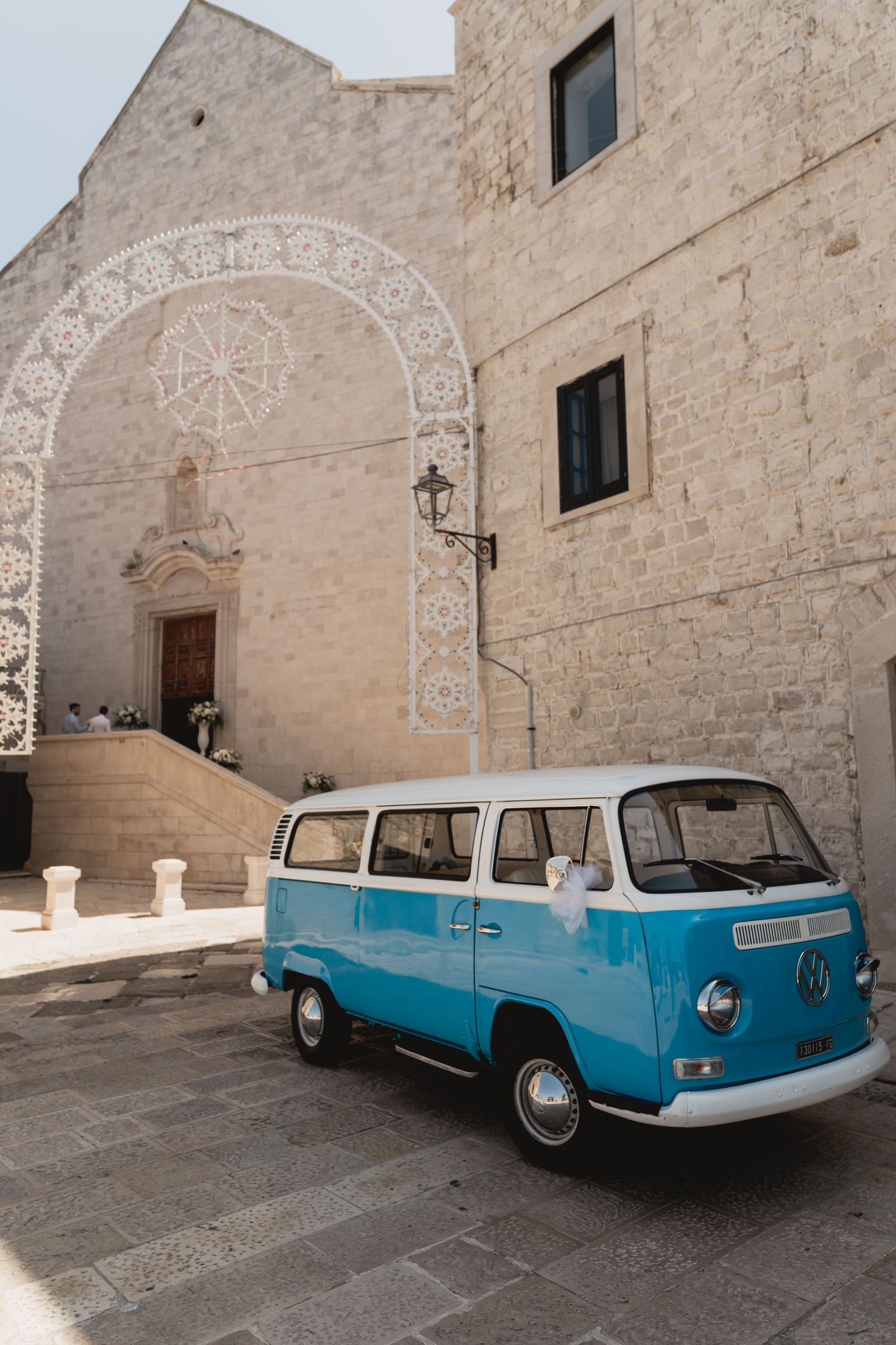 Classic blue and white VW van parked in front of a historic stone church with ornate architectural details.