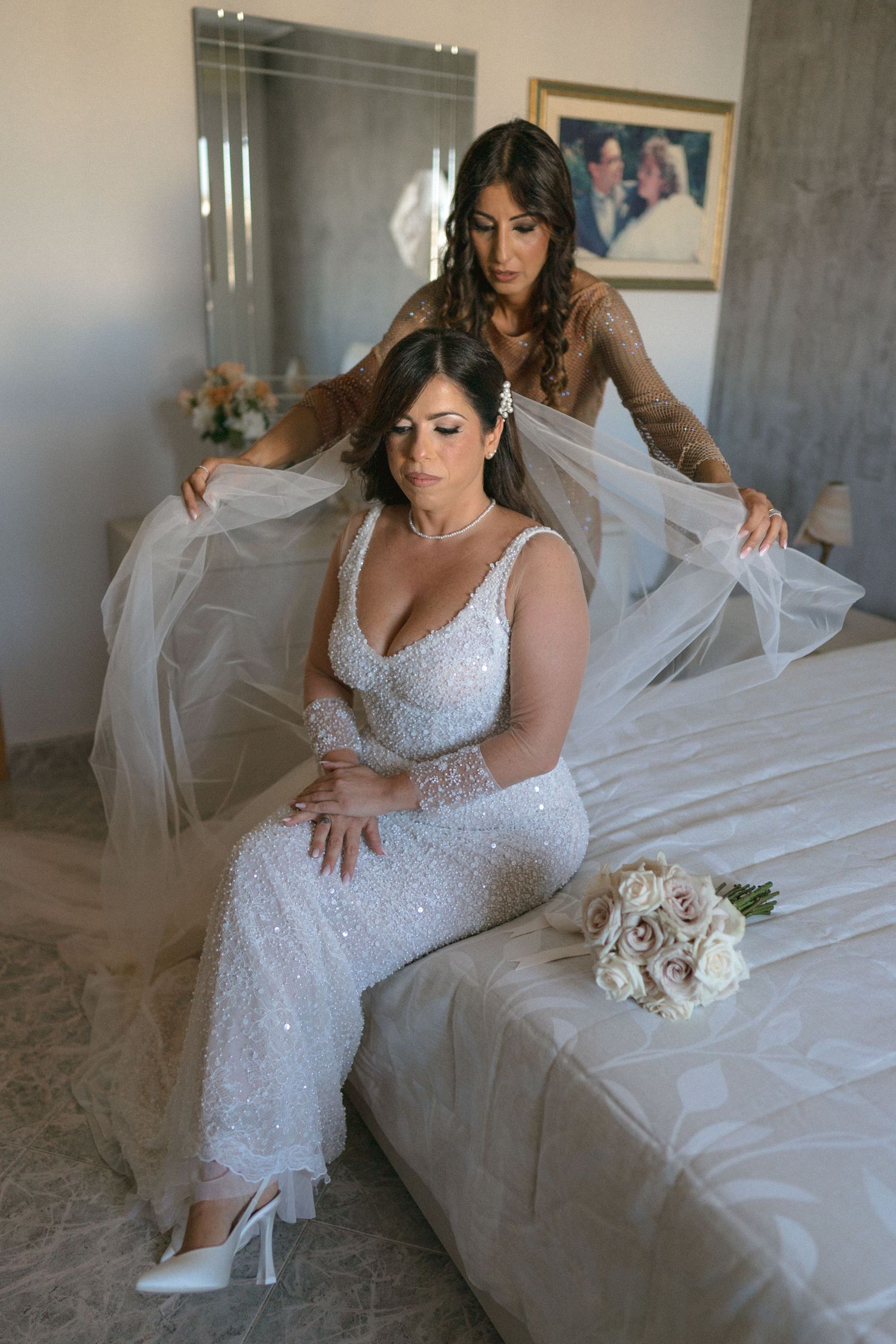 A bride in a sparkly white dress sits while her veil is being adjusted.