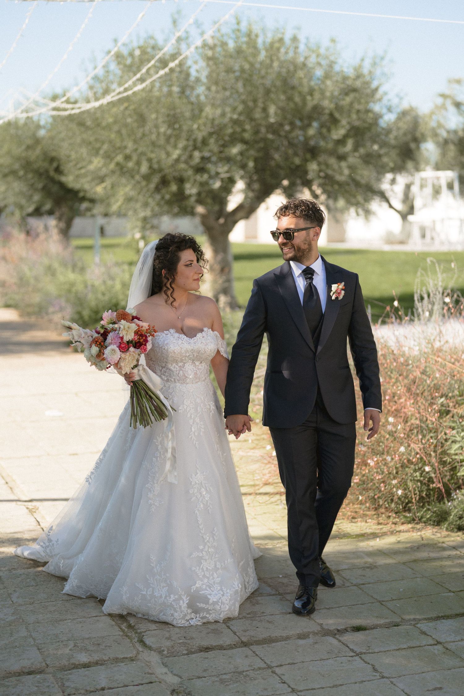 Bride and groom walking hand in hand through an olive tree garden on their wedding day.