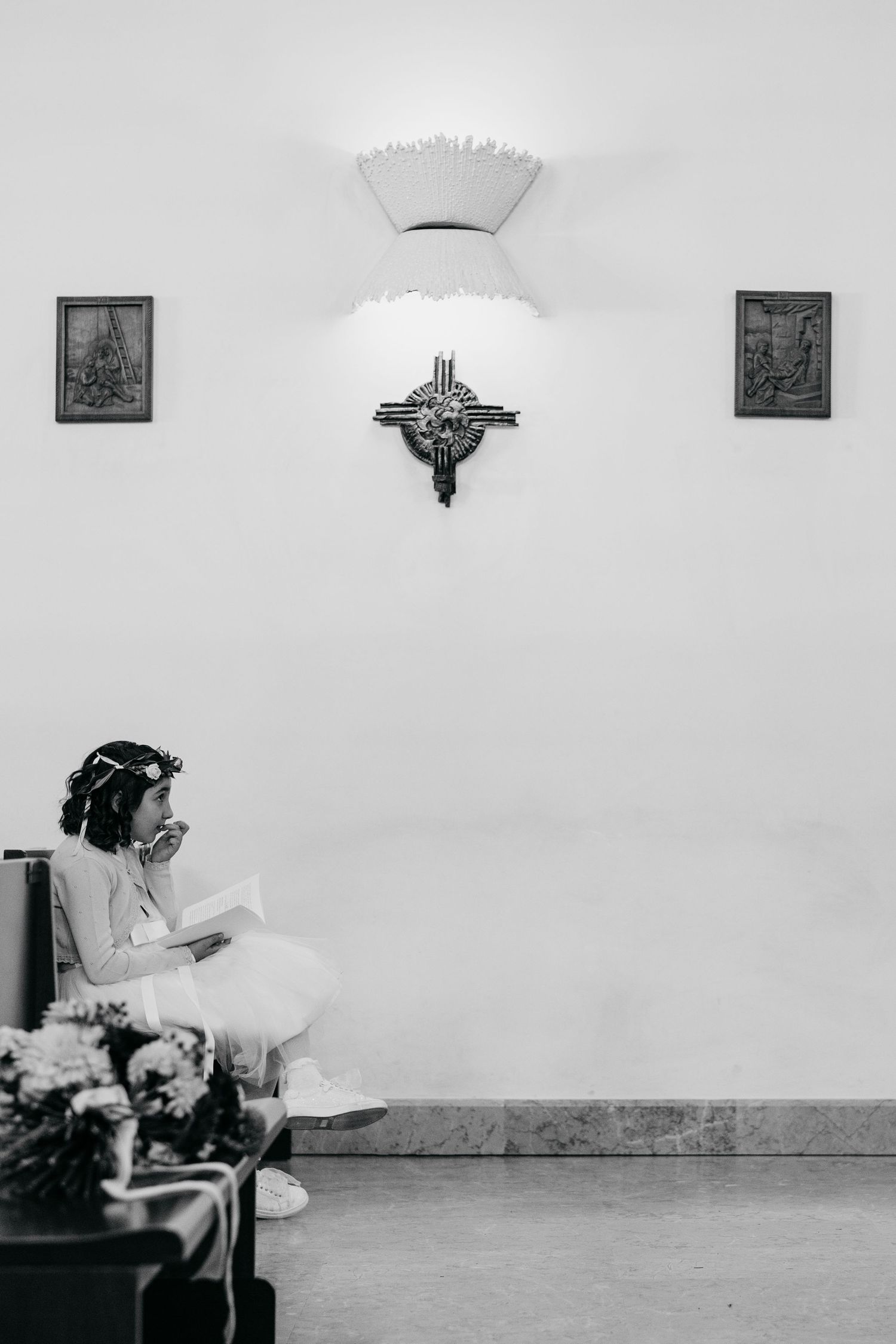A young girl in a white dress and floral crown sits alone in a church reading, with a cross on the wall behind her.
