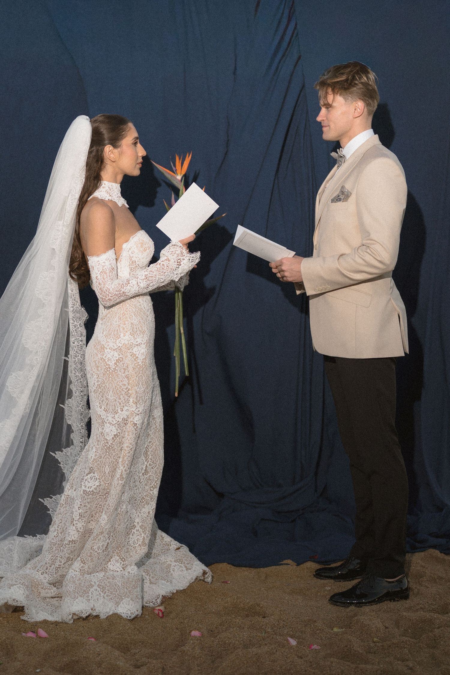 Bride in a lace wedding dress and veil holds a bird of paradise flower and reads vows facing a groom in a beige blazer.