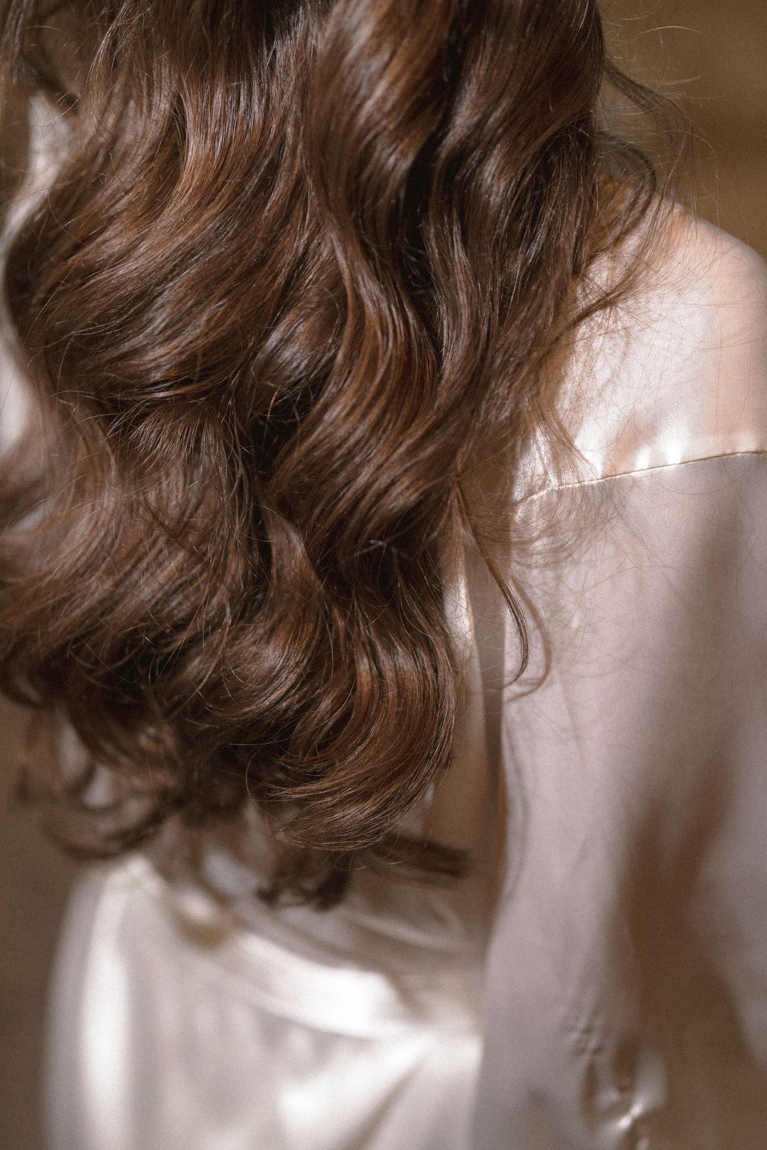 Long, glossy brunette waves fall over the back of a woman wearing a soft pink satin robe in warm, golden light.